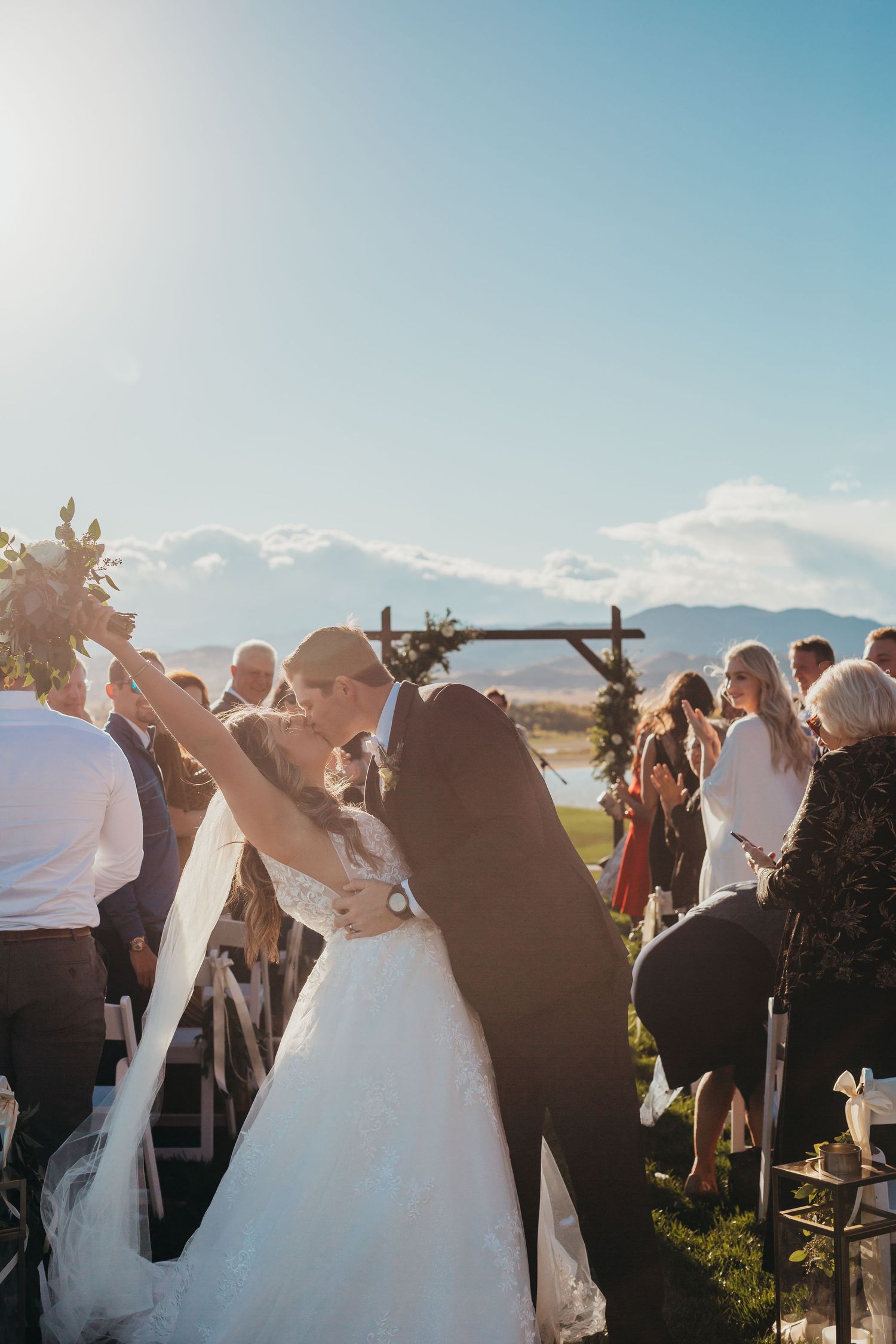 Newlyweds celebrate during an outdoor mountain ceremony with guests and floral arch.