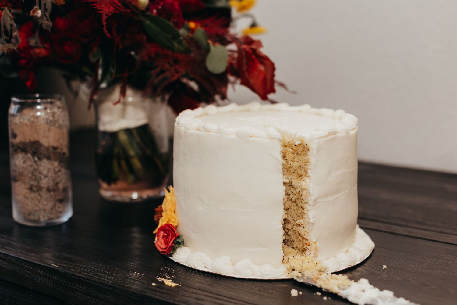 A white frosted layer cake with crumbs visible on a dark wooden table next to red flowers in a vase.