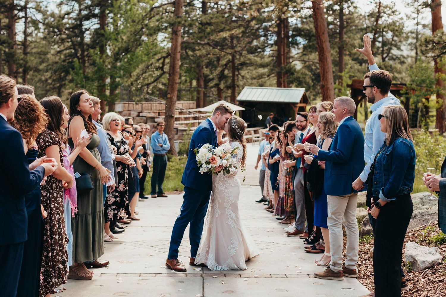 Wedding guests line a stone pathway in a pine forest as a couple shares their first kiss as newlyweds.