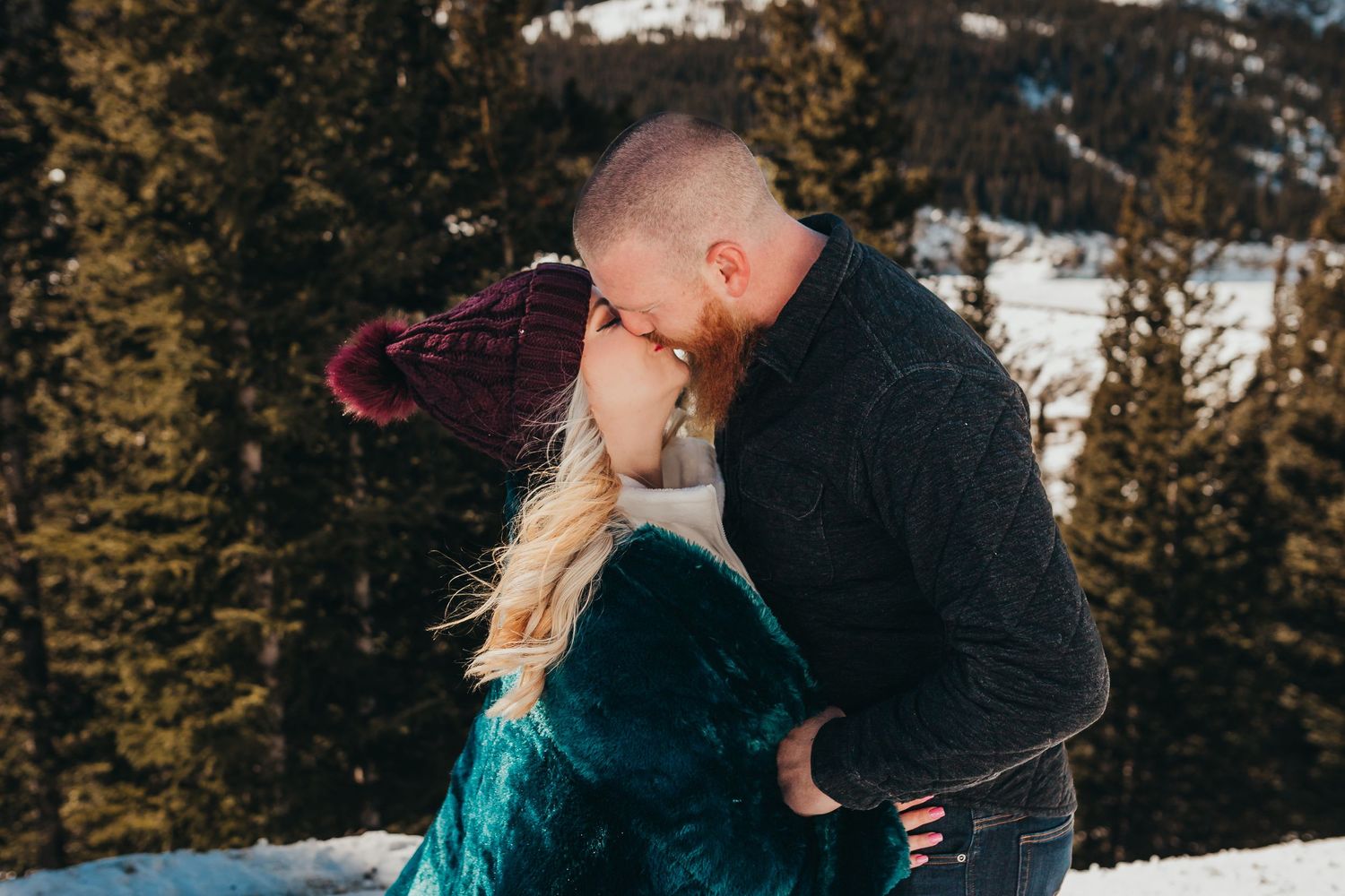 Couple embraces in winter scene with snow-covered mountains and evergreen trees.
