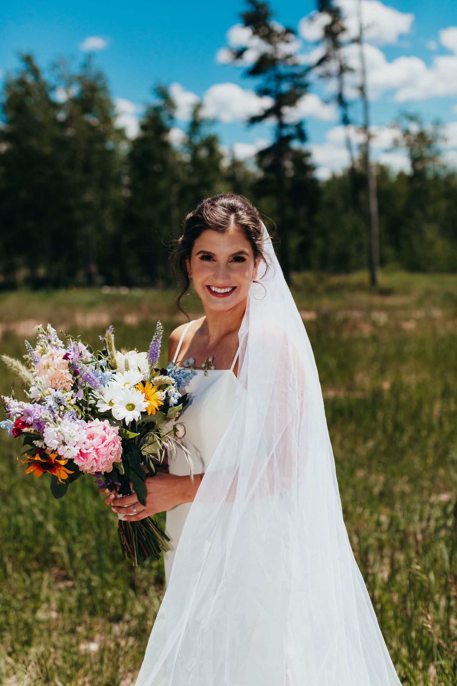 A bride holds a colorful wildflower bouquet while standing in a mountain meadow.