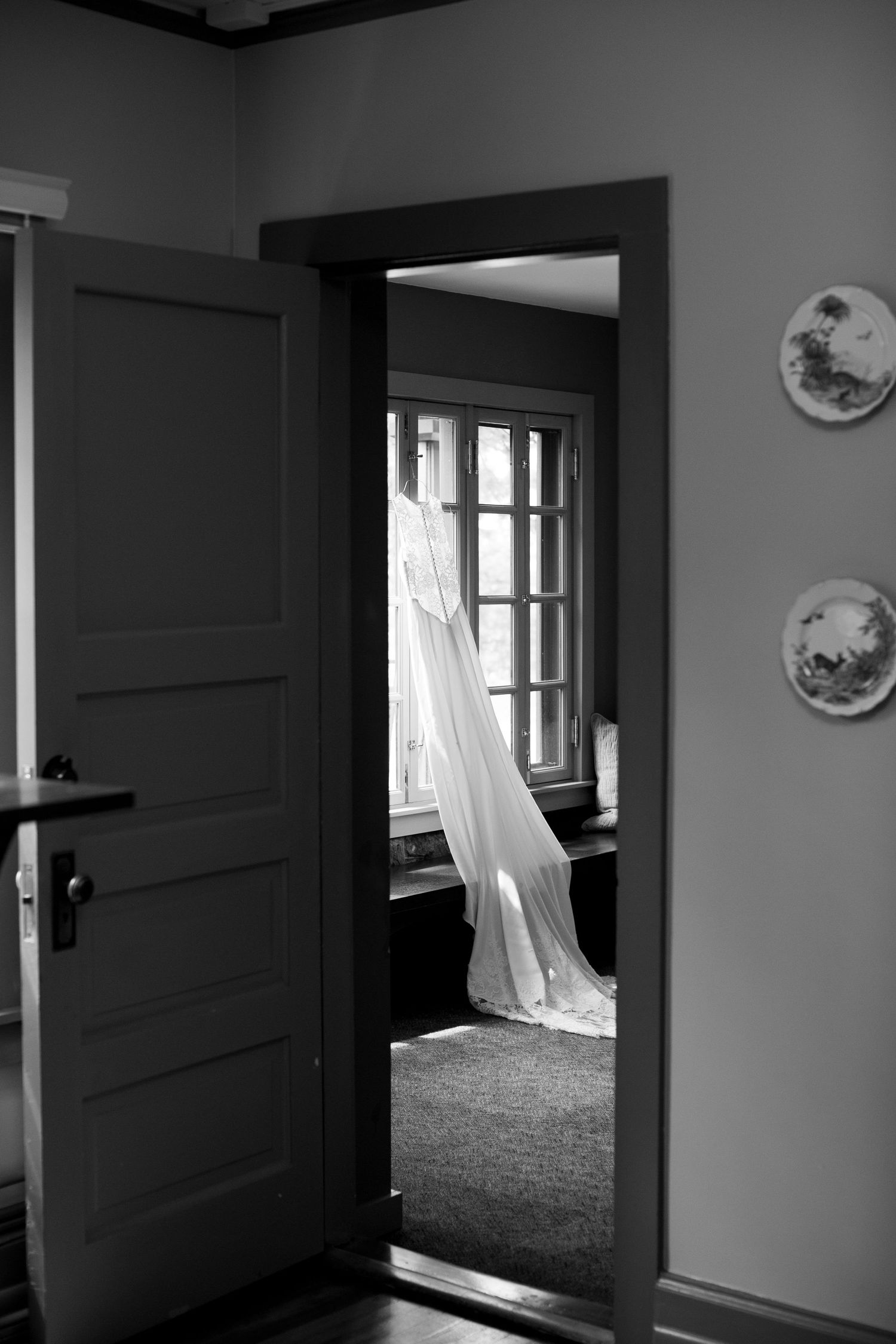 Black and white photo of wedding dress hanging in window frame seen through doorway.