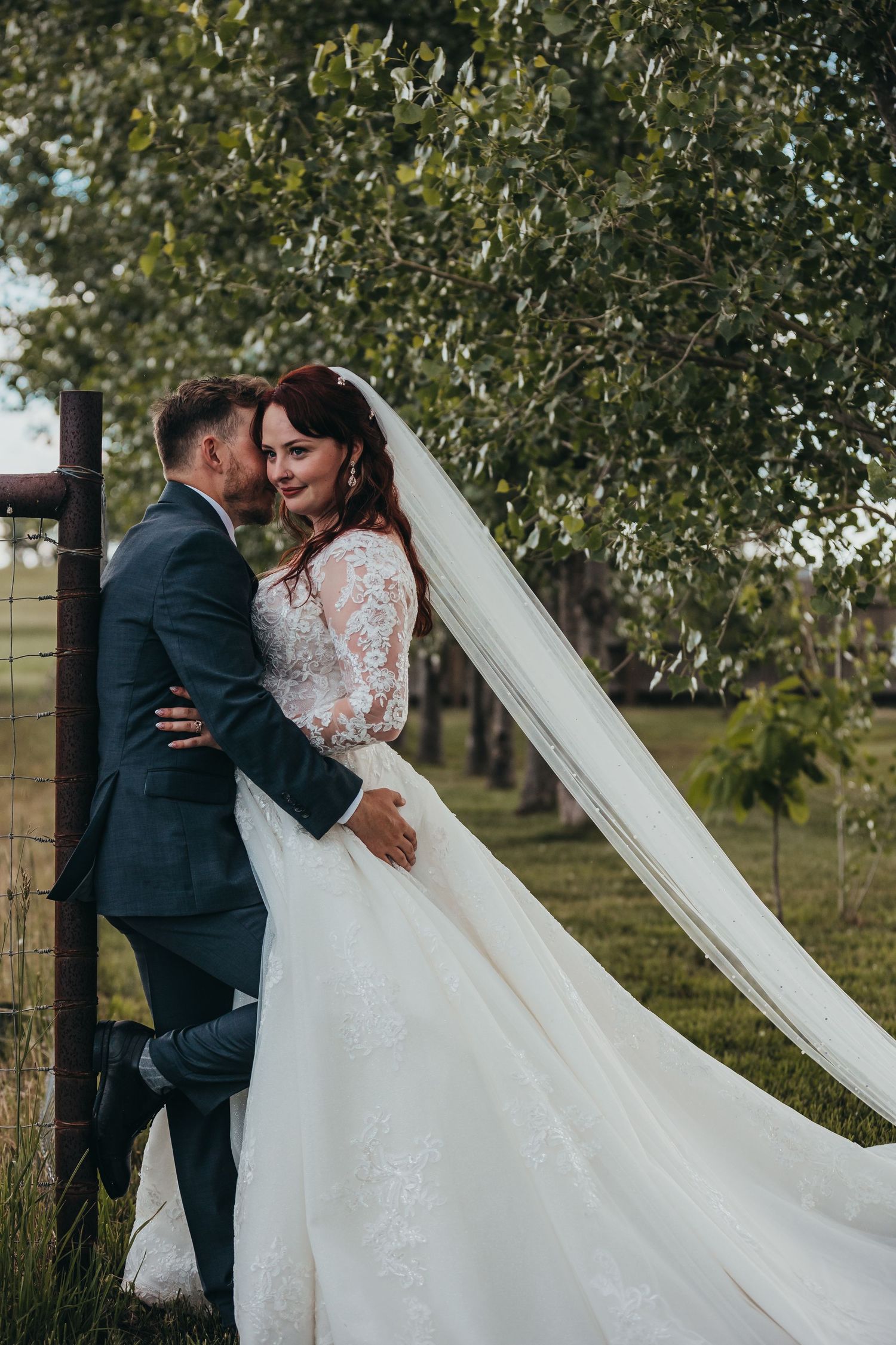 Romantic outdoor wedding portrait with flowing veil and lace dress against rustic fence.