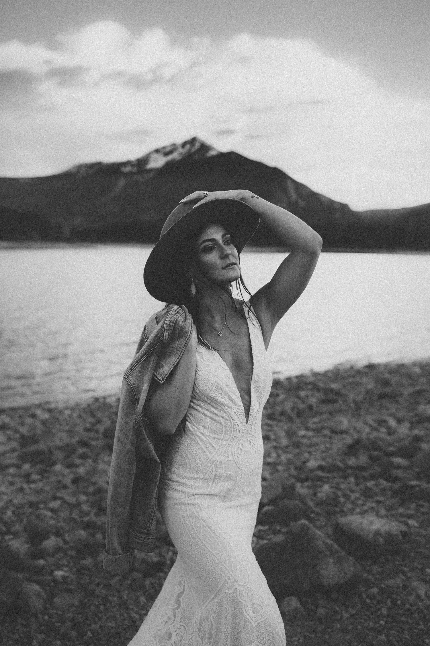 Bride in plunging white dress and hat poses against mountain lake backdrop in black and white photograph.