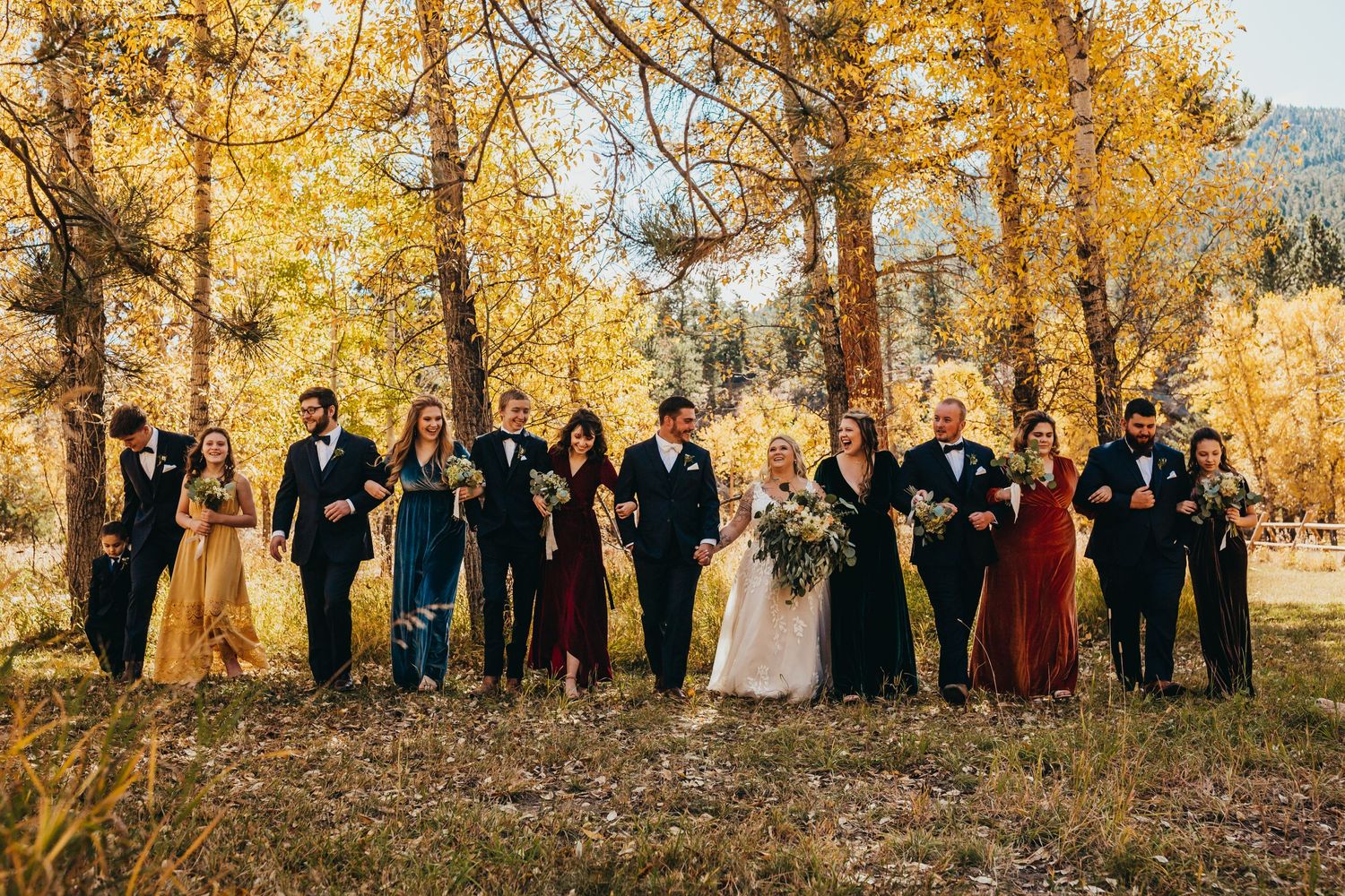 Large wedding party in colorful attire poses together among golden fall trees.