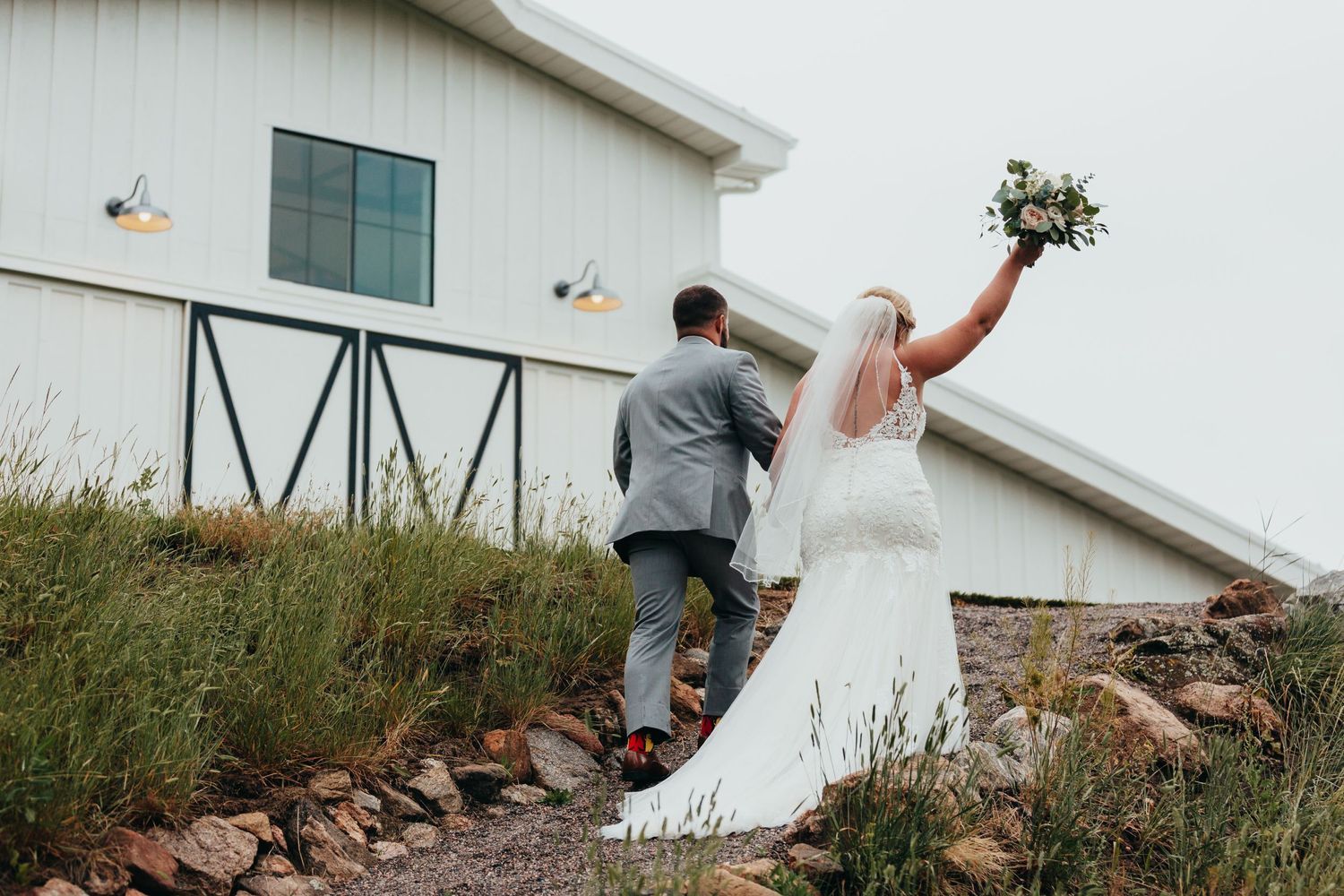 A couple climbs up a grassy hill next to a white building during their wedding.