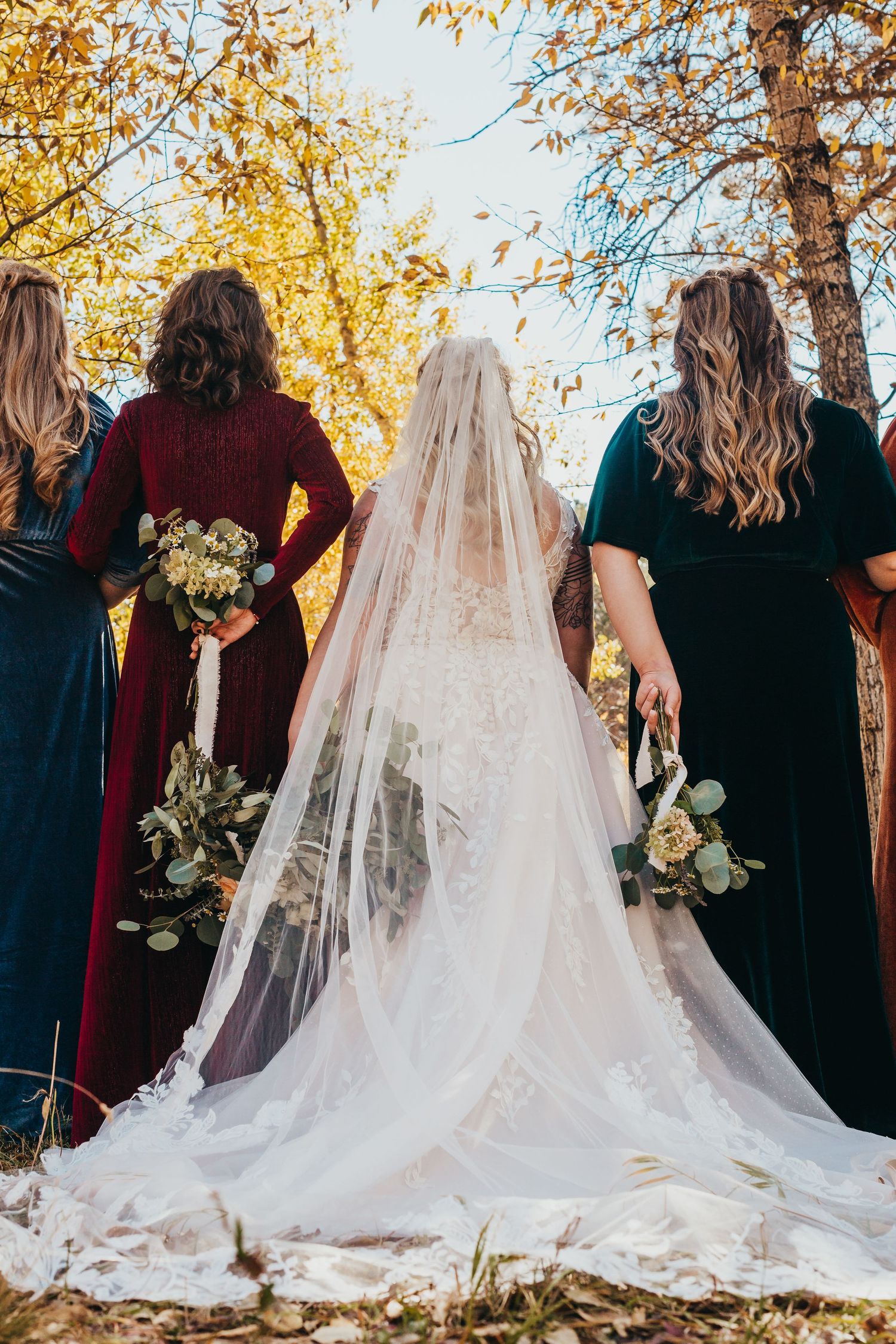 Three bridesmaids in jewel-toned dresses stand with the bride's flowing veil against fall foliage.