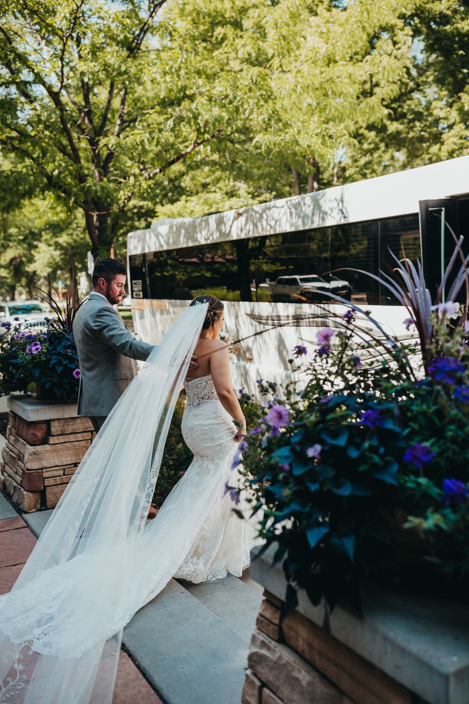Bride with flowing veil walks near decorative planters with purple flowers by white bus.