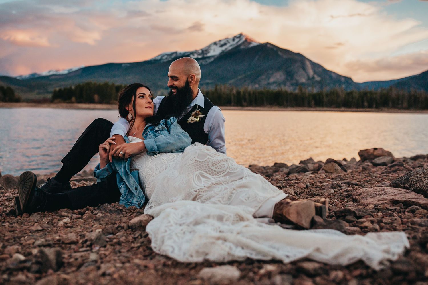 Couple shares intimate moment on rocky lakeshore with mountain backdrop at sunset.