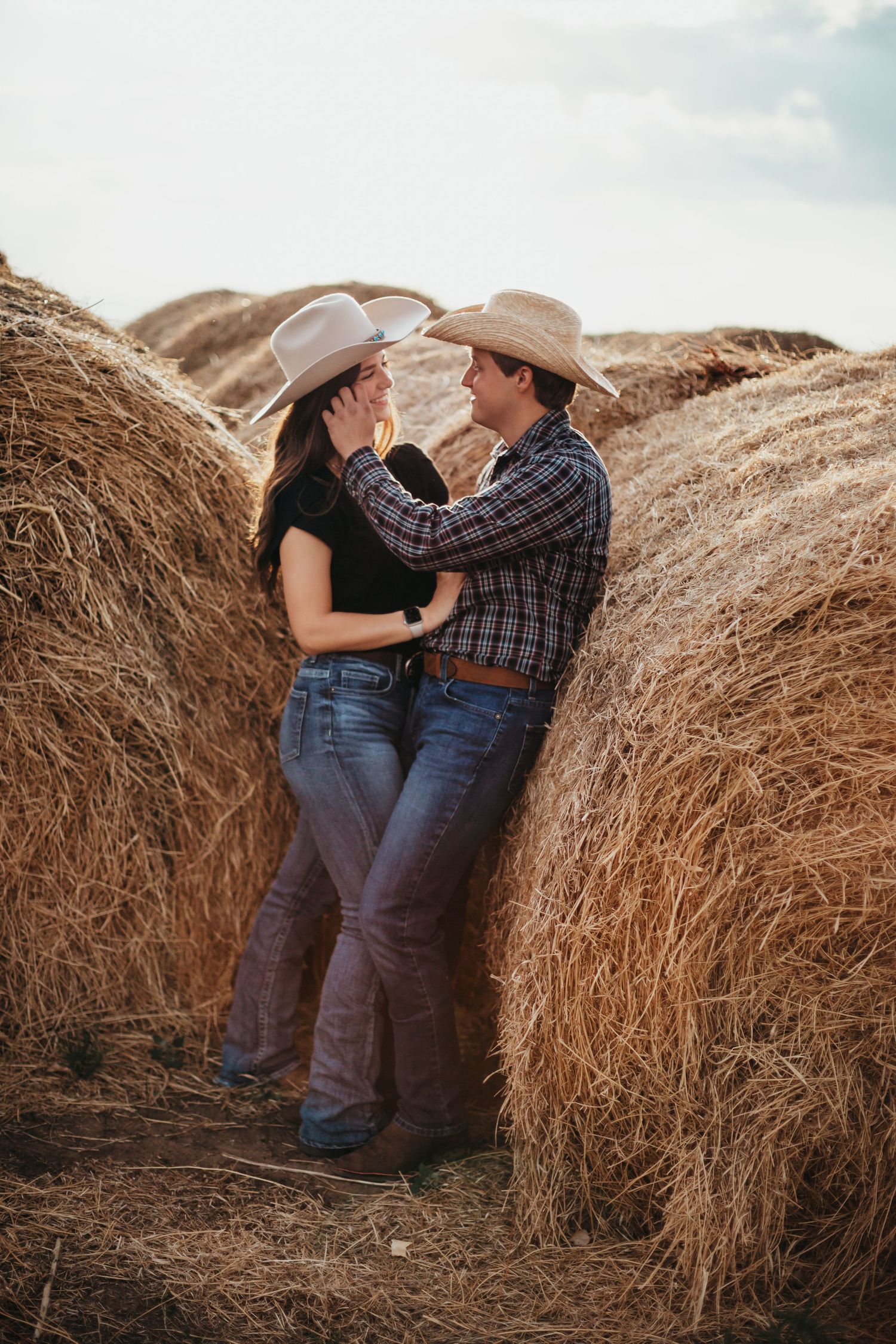 Romantic couple in western attire embracing against hay bales at sunset.