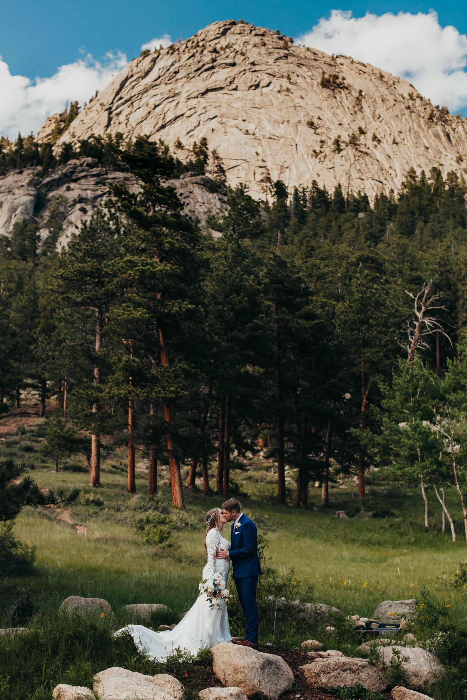 Wedding couple standing before dramatic mountain landscape with pine trees.