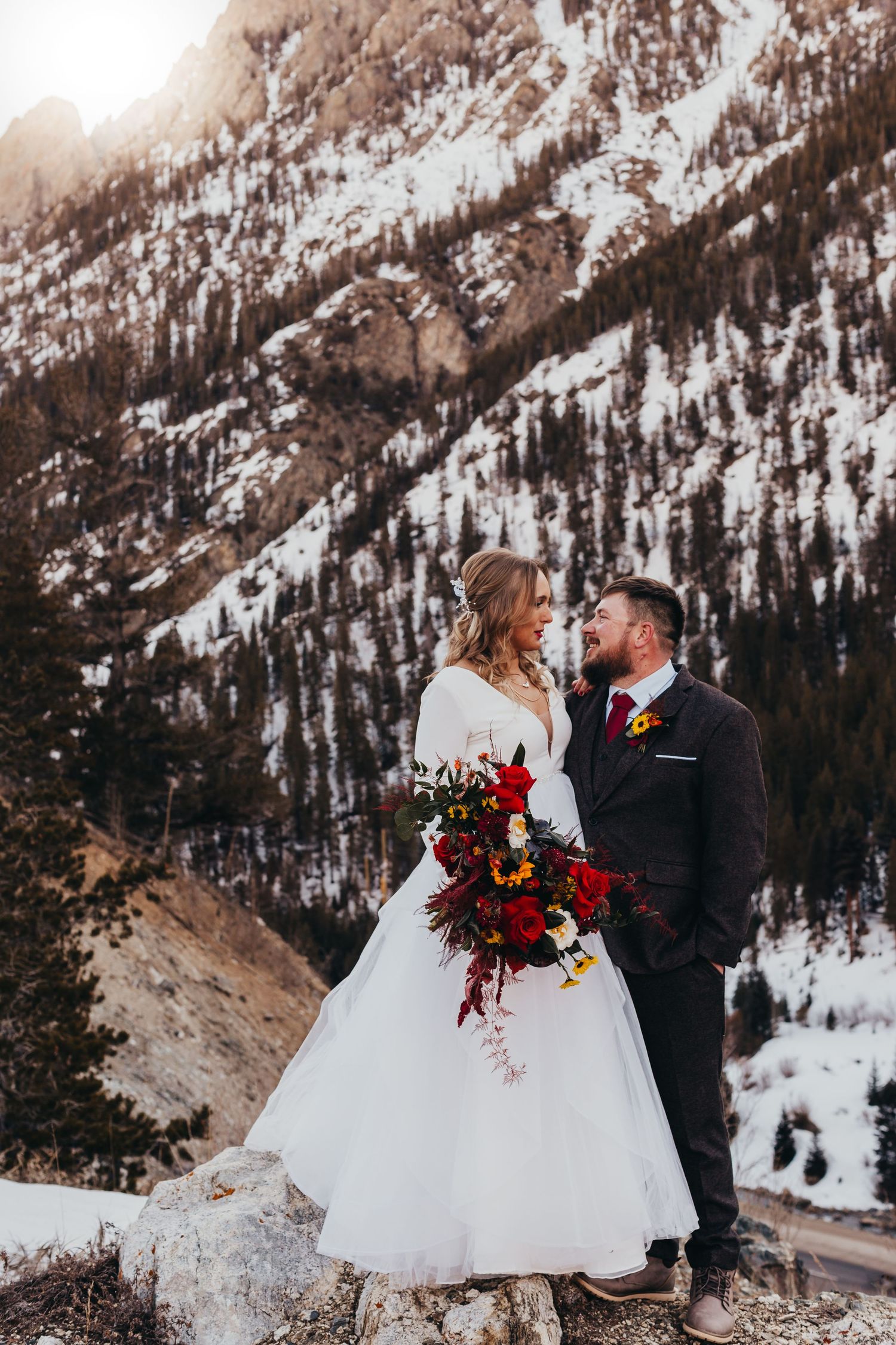 A romantic winter wedding photo against a snow-covered mountain backdrop with vibrant red bouquet.