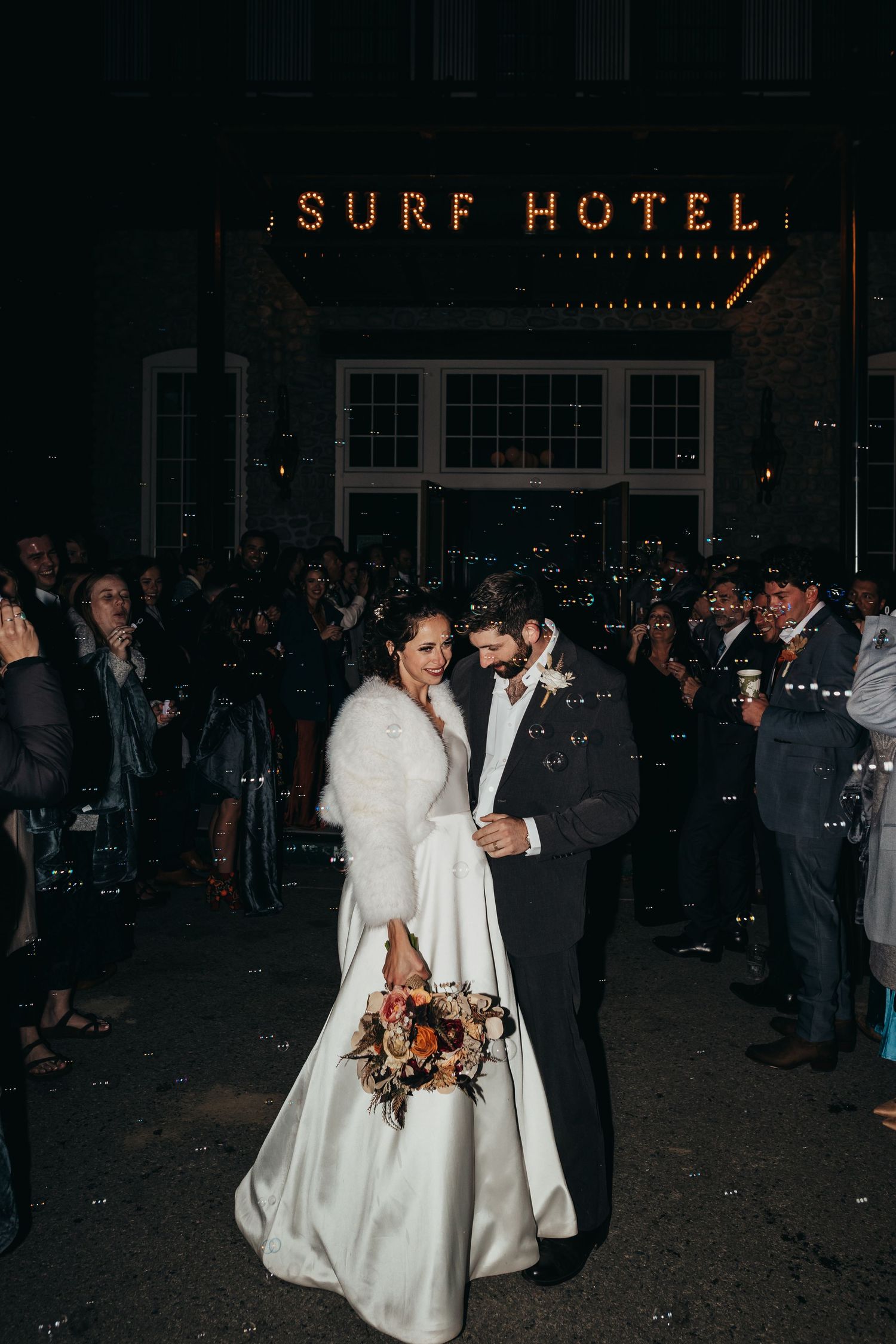 Newlyweds pose beneath the vintage Surf Hotel sign at night surrounded by wedding guests.