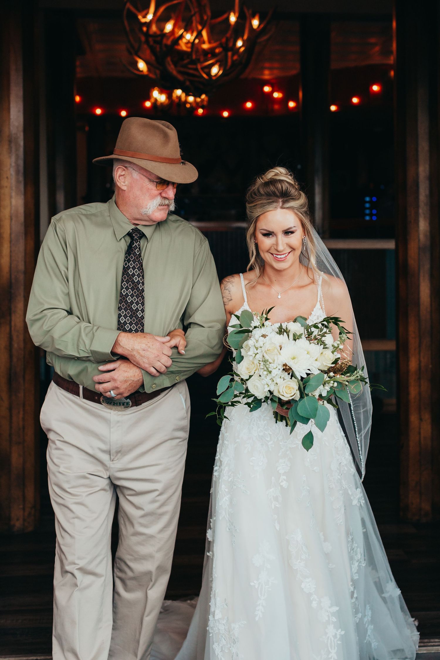 A bride in a white dress holding a floral bouquet walks arm in arm with a figure in a tan hat and green shirt.