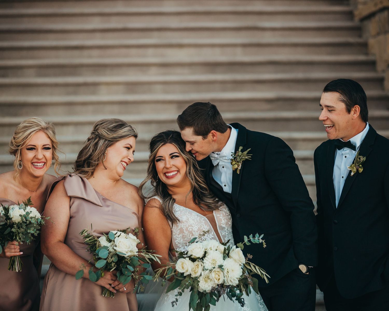 Wedding party members share a candid laughing moment on stone steps.