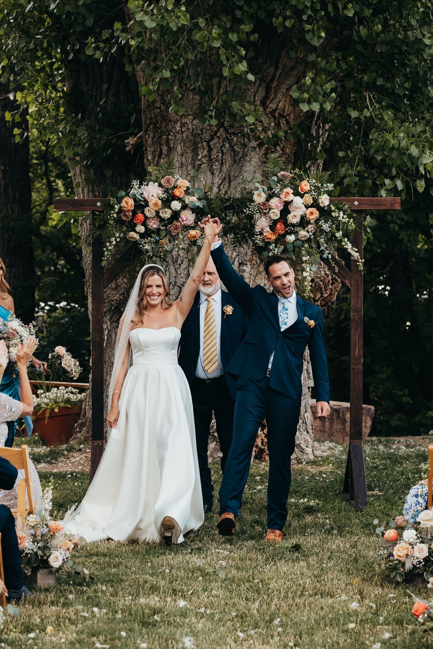 Newlyweds celebrate during outdoor garden wedding ceremony under floral arch with peach and white flower arrangements.