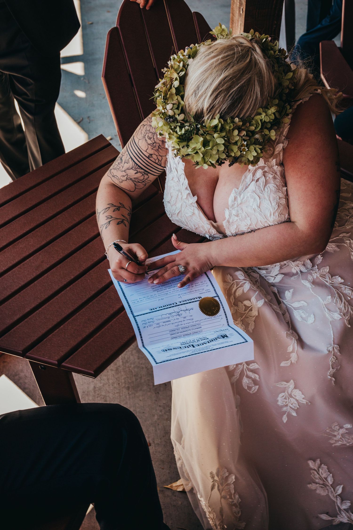 Person in lace dress and flower crown signing wedding documents at wooden table.