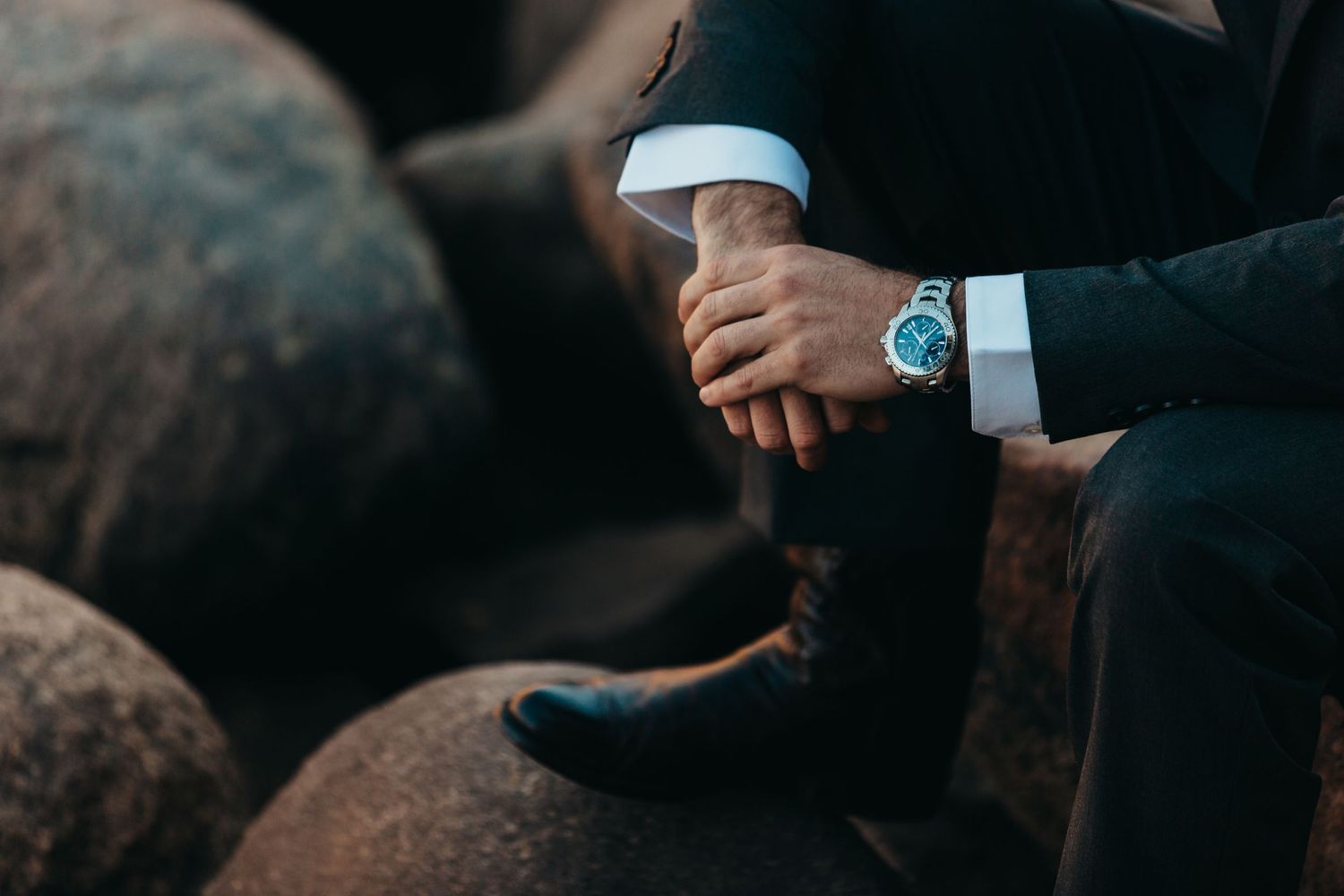 Close-up shot of a formal suit sleeve with a blue watch on the wrist against dark leather furniture.