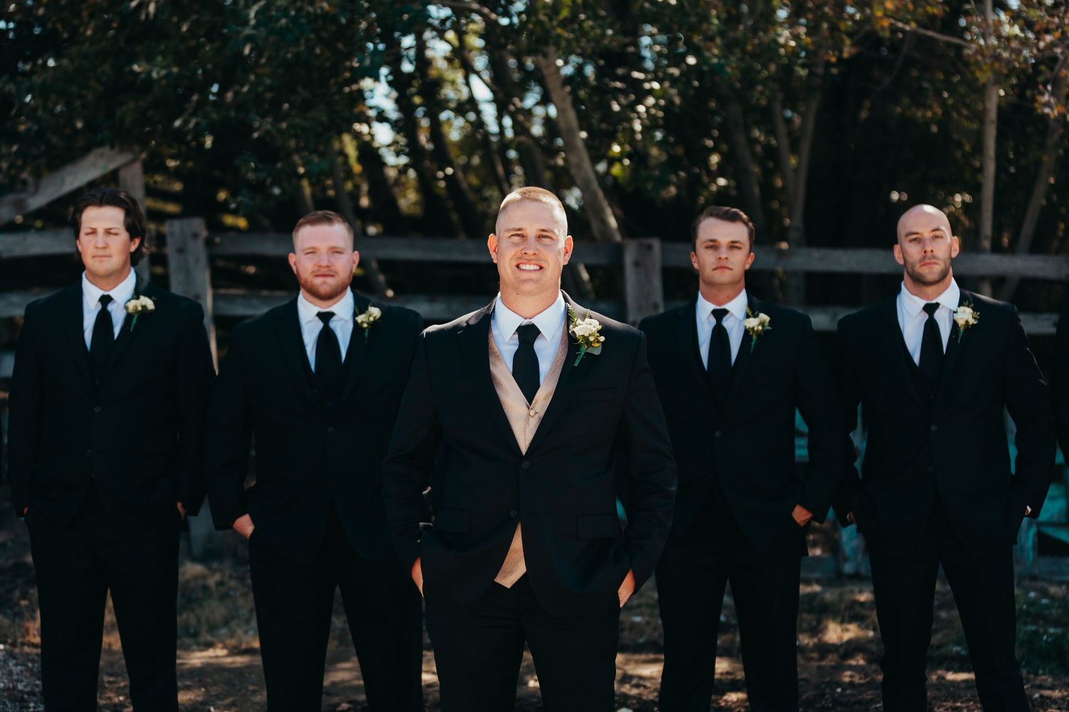 Groomsmen in black suits stand with groom in tan suit outdoors at wedding.