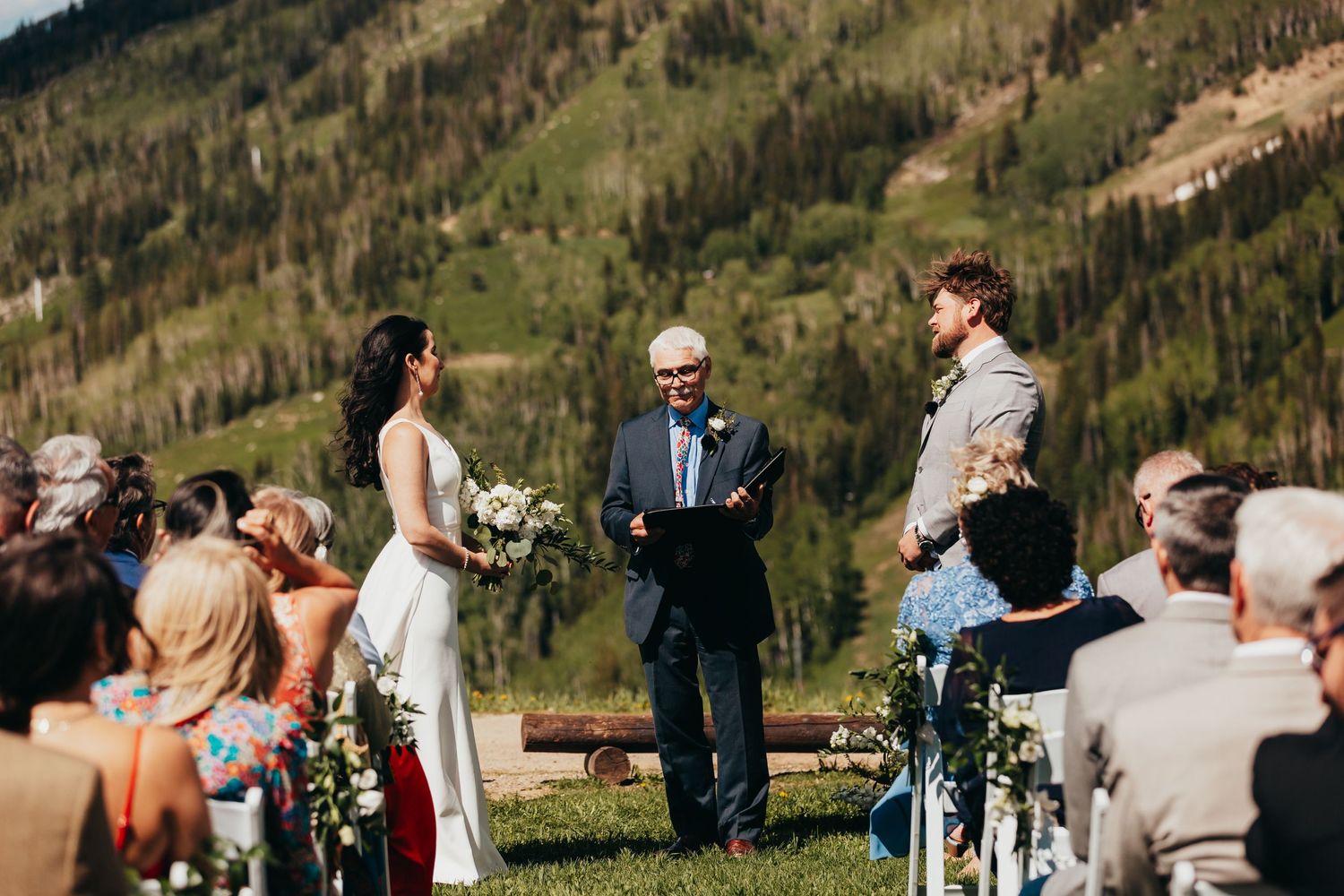 An outdoor mountain wedding ceremony with white chairs set up on a grassy slope.