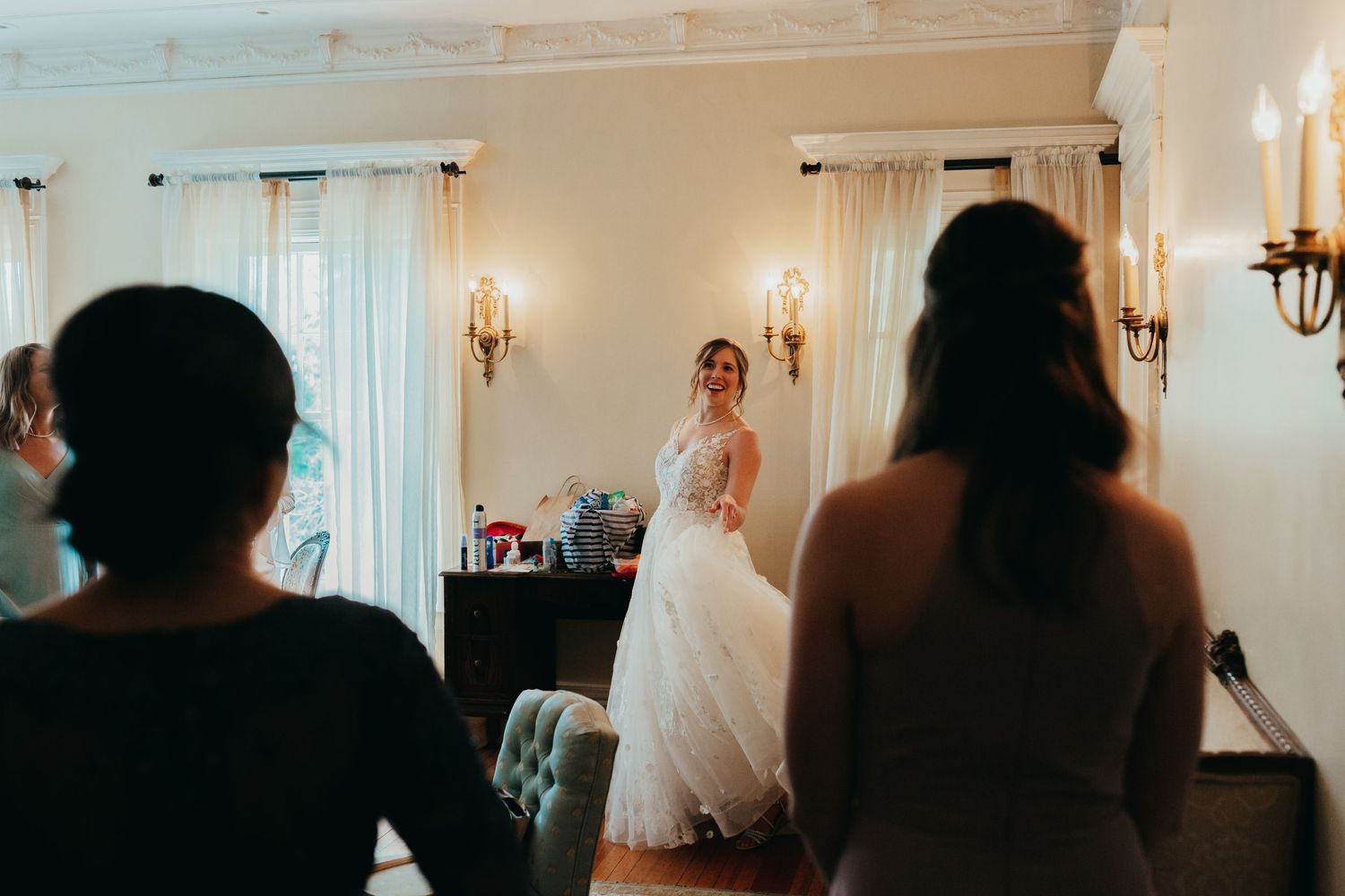 A bride in a flowing white gown stands in an elegant room with wall sconces and white curtains.
