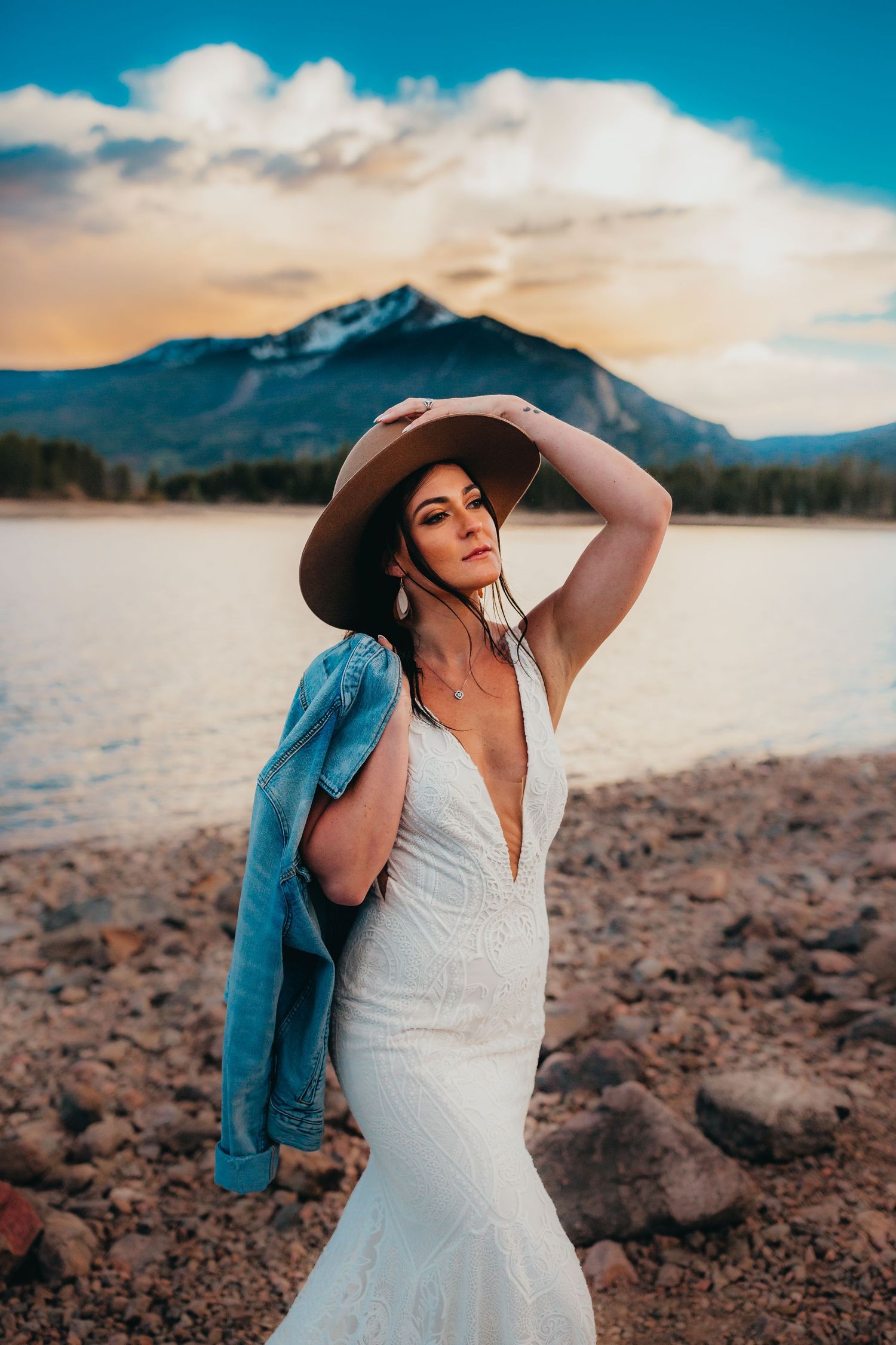 Bride in mermaid gown with denim jacket strikes dramatic pose by mountain lake at sunset.