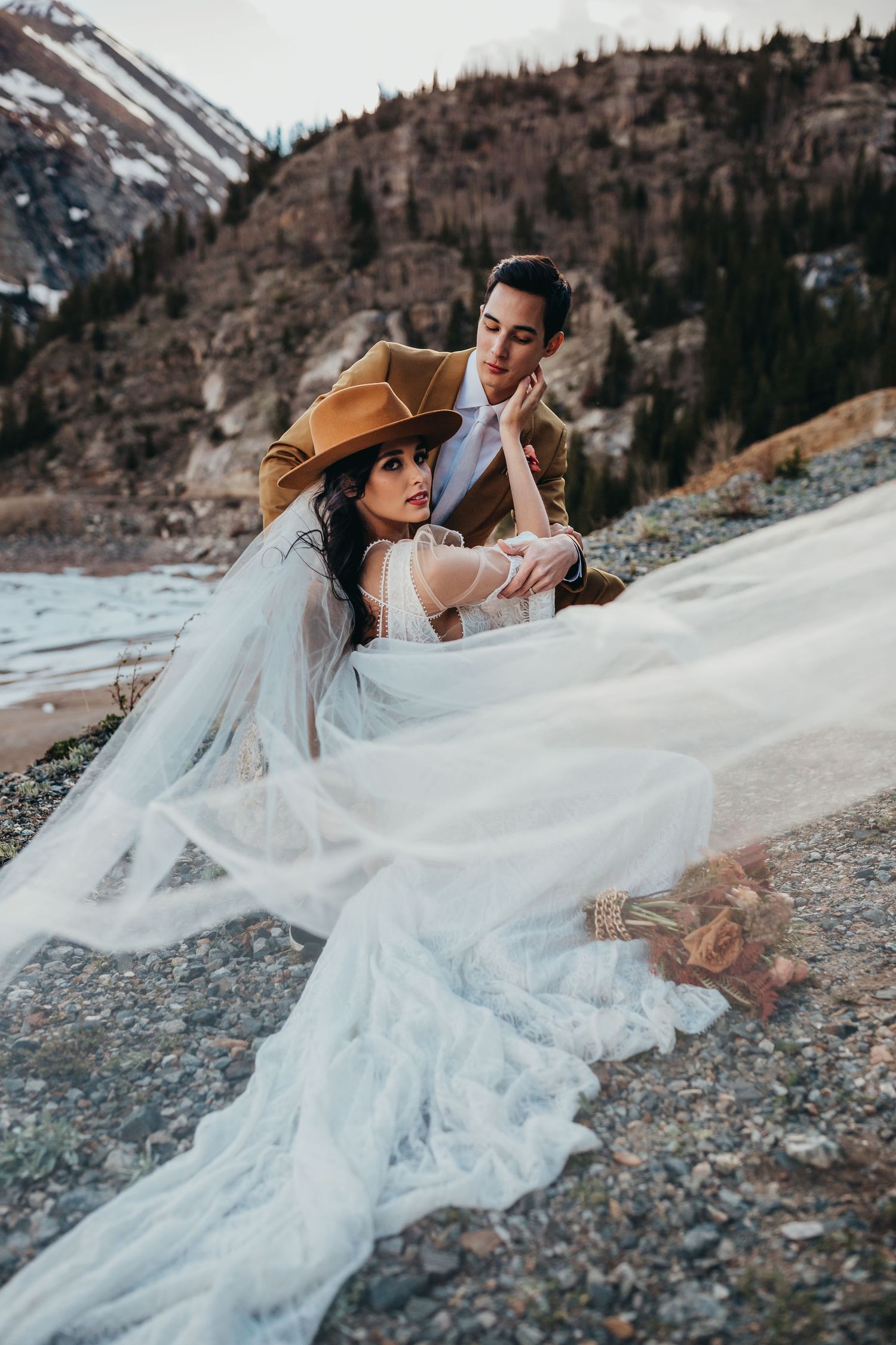 Wedding couple poses dramatically on mountain terrain with bride's veil floating in the breeze.