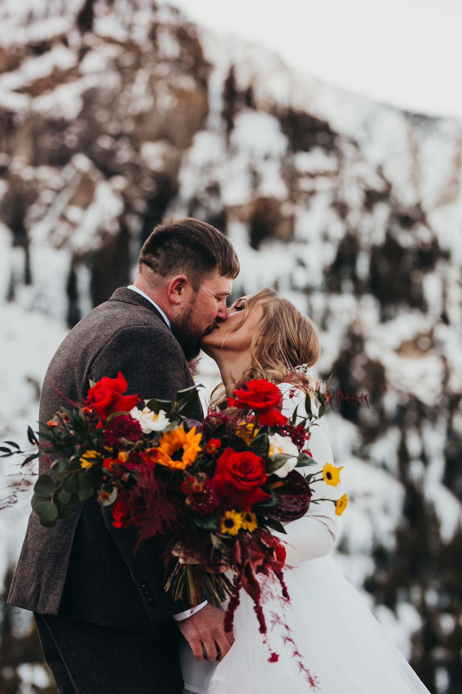A winter wedding kiss captured against snow-covered mountains with colorful floral bouquet.