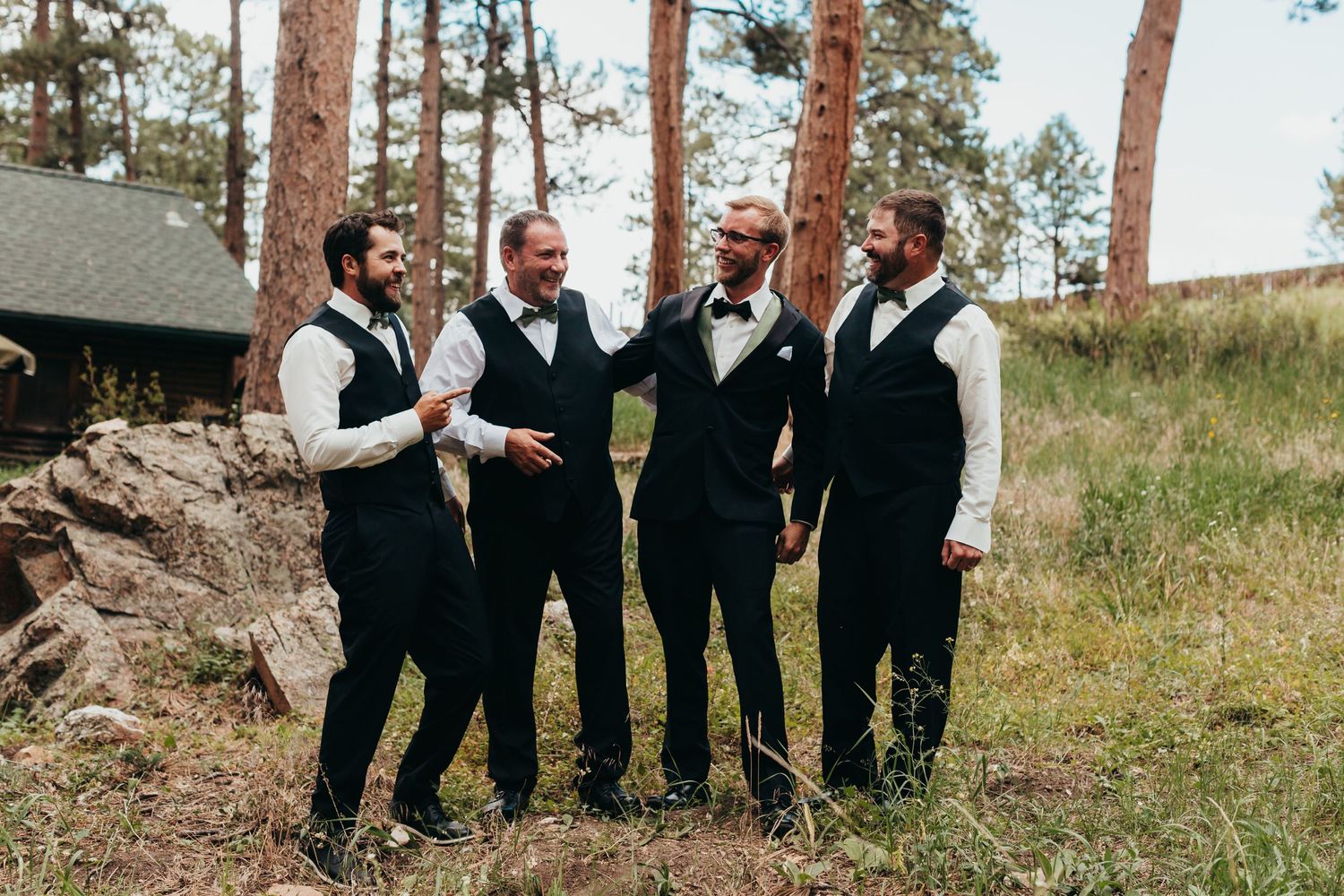 Groomsmen in black vests and bow ties standing together outdoors among tall pine trees.