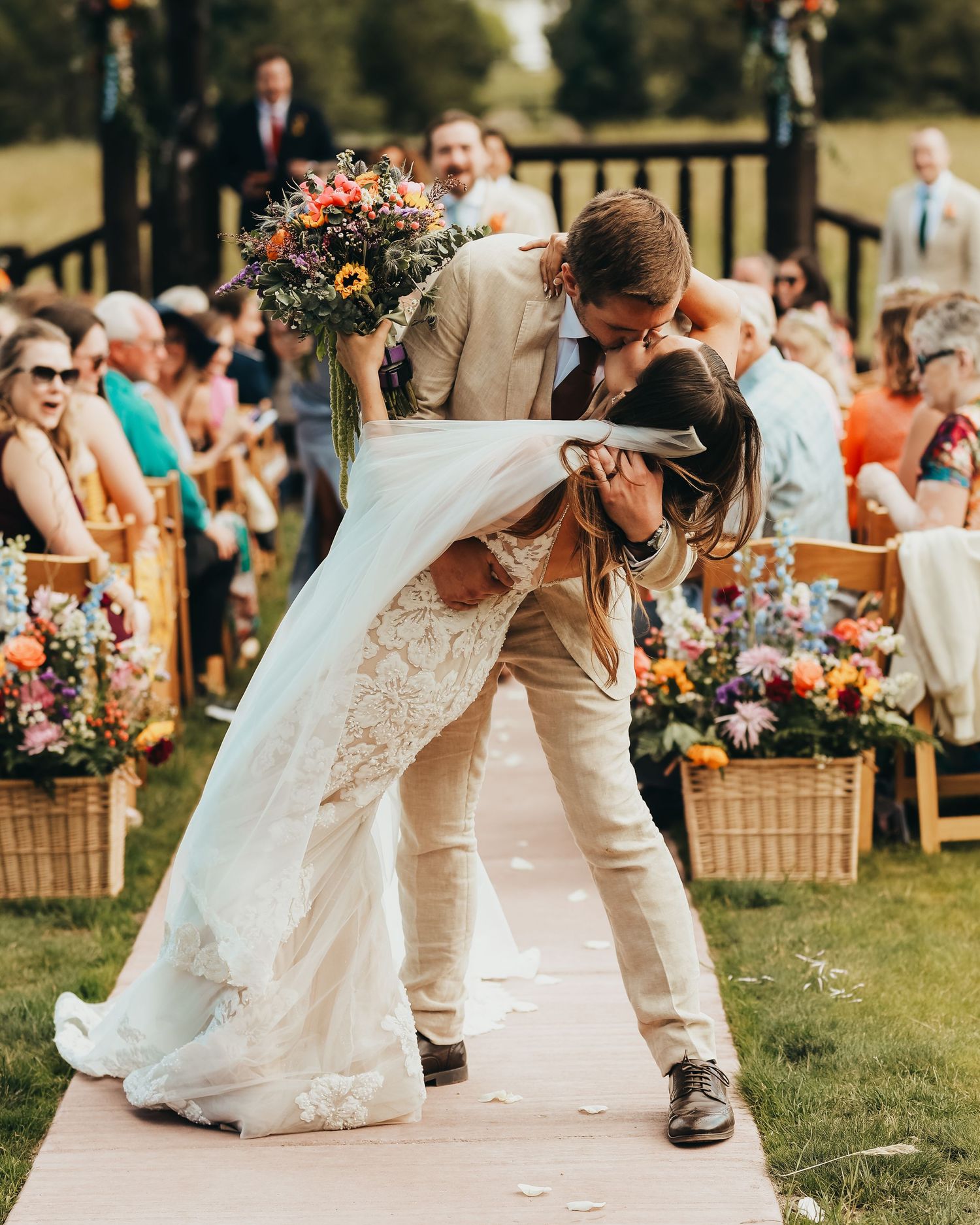 Romantic wedding dip kiss on an outdoor aisle decorated with colorful floral arrangements.