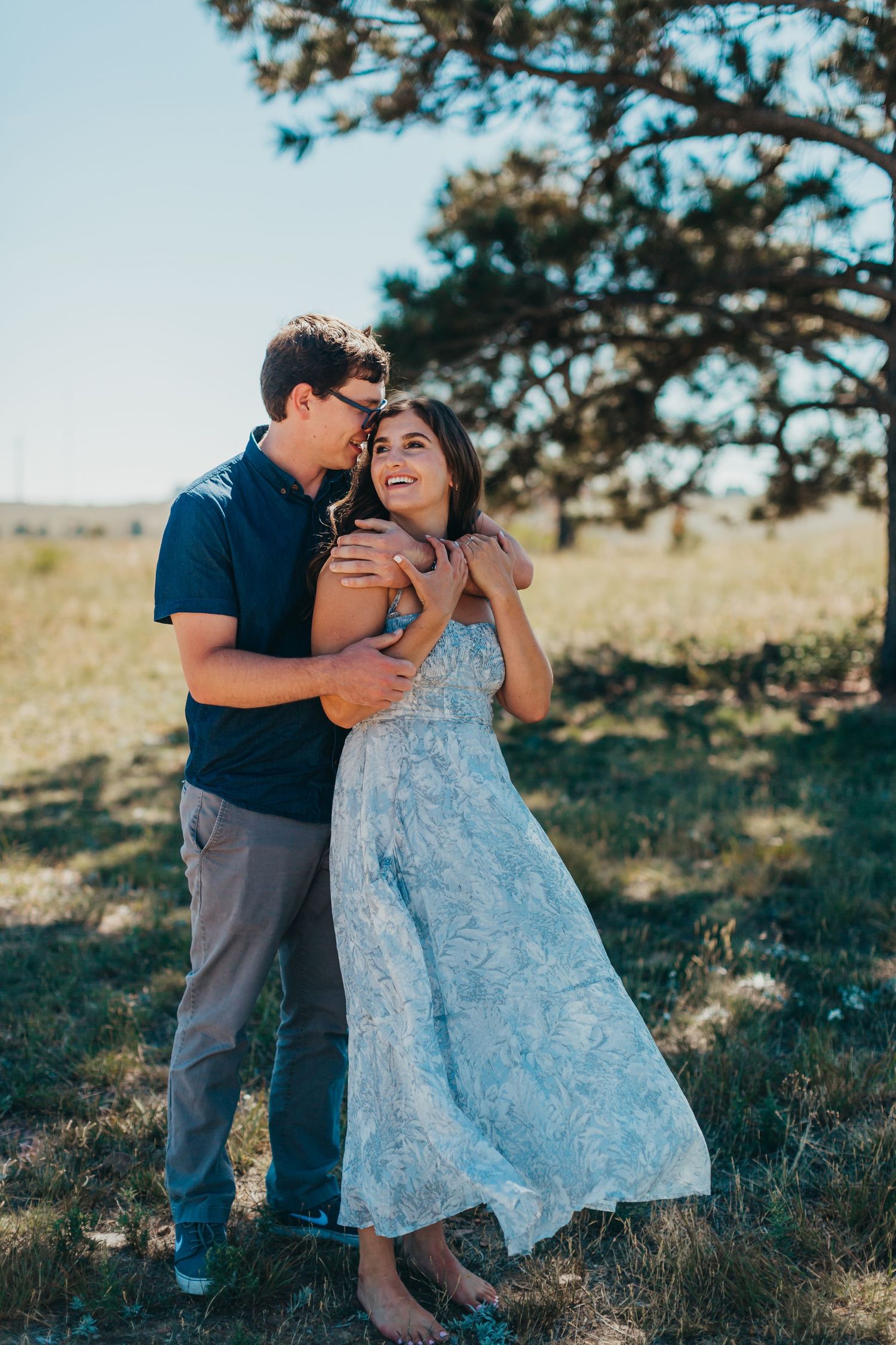Romantic couple embracing in sunlit meadow with pine tree in background.