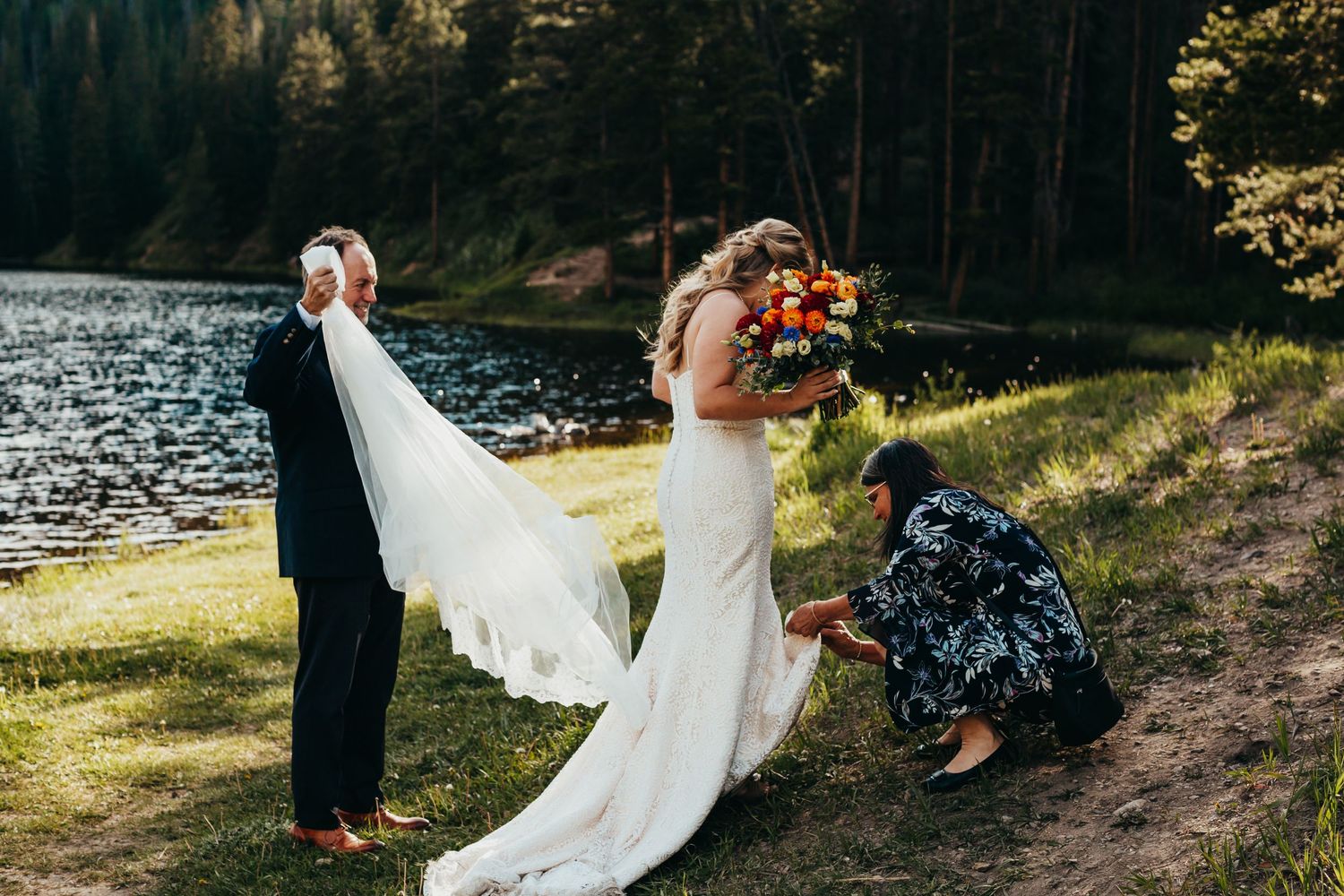 Wedding party adjusting bride's veil by lakeside in forest setting.