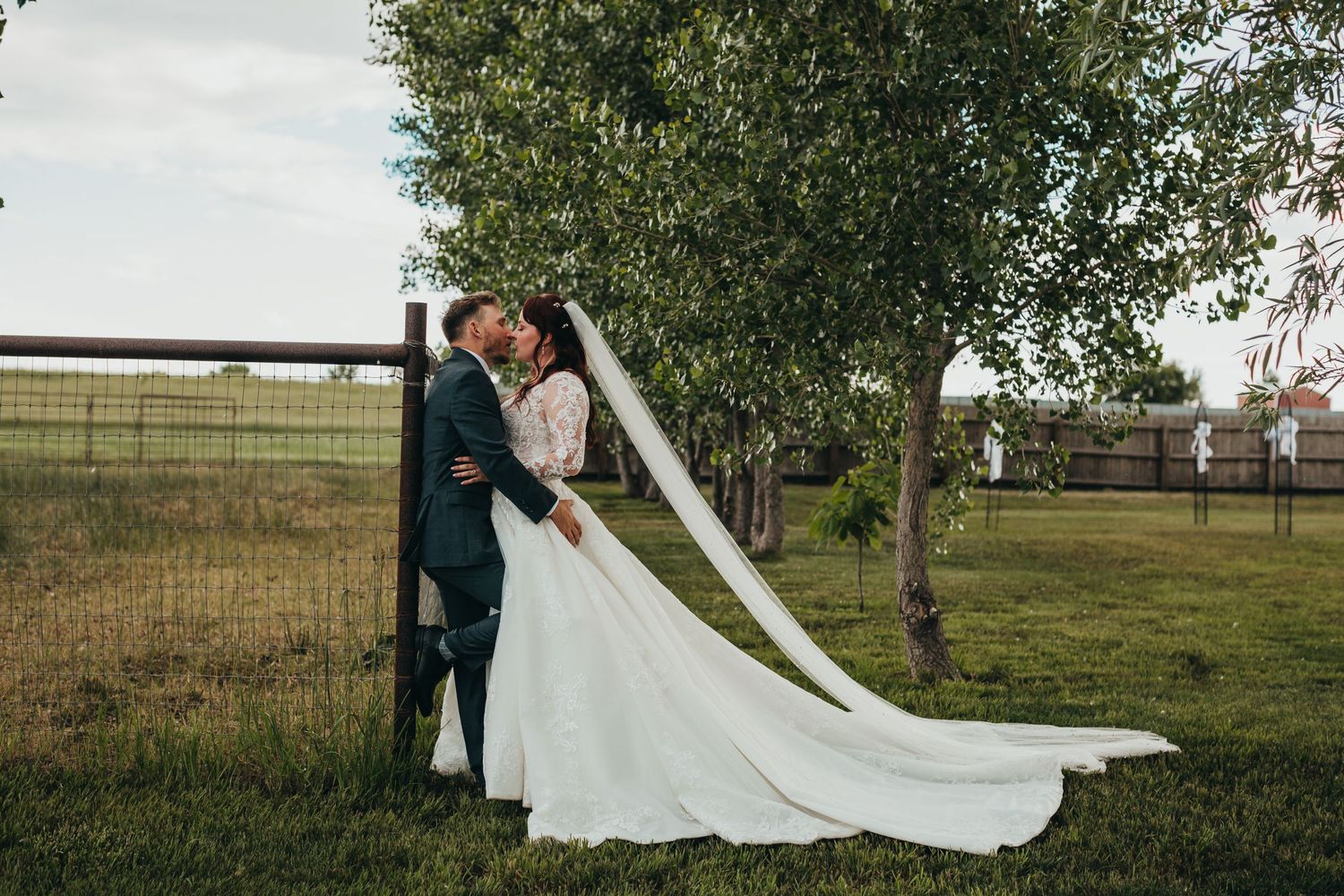 A romantic wedding couple embraces against a pastoral fence backdrop with a flowing white dress train.
