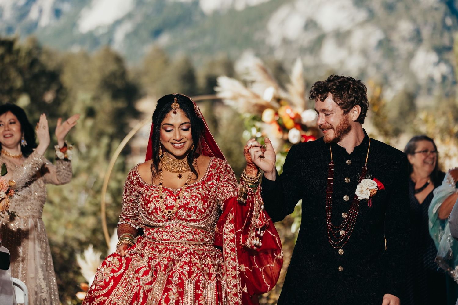 Outdoor mountain wedding celebration with couple in traditional red and black attire.