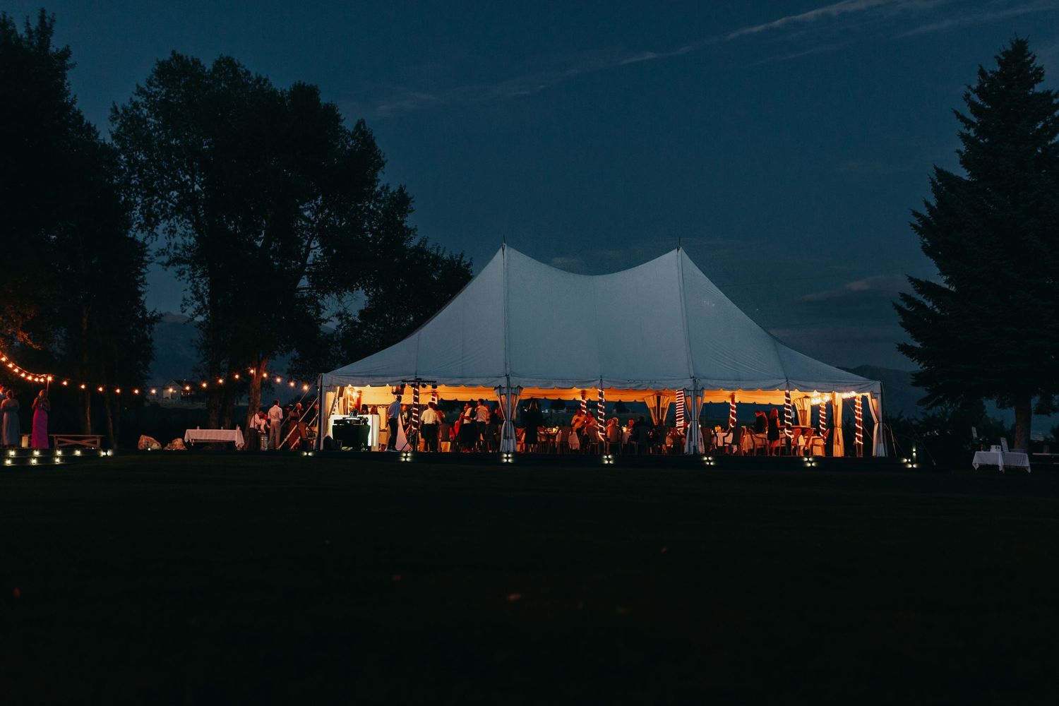 Large white wedding tent illuminated at night with string lights and guests celebrating inside.