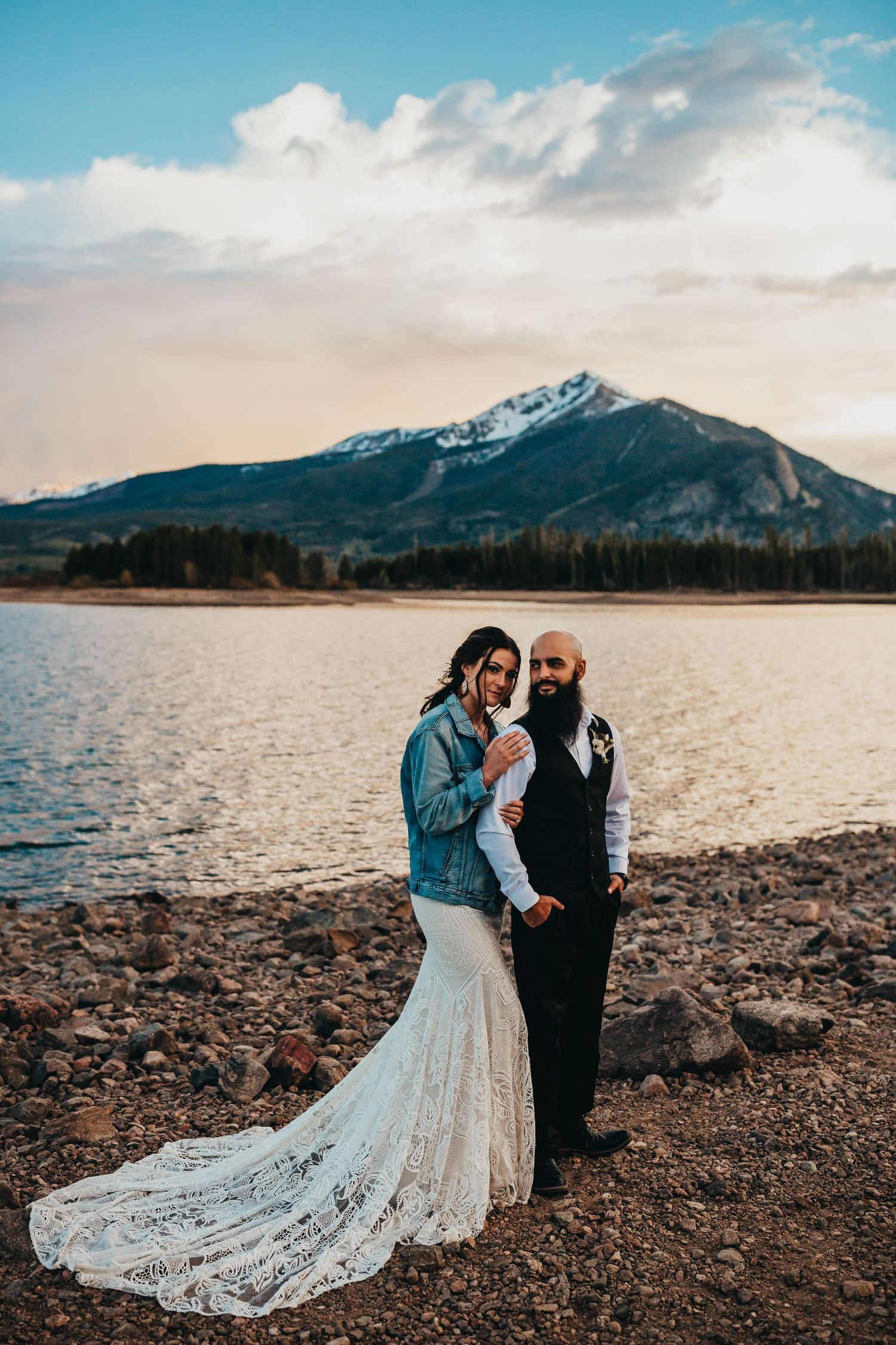 Stunning lakeside wedding portrait with snow-capped mountain peak and dramatic sky.