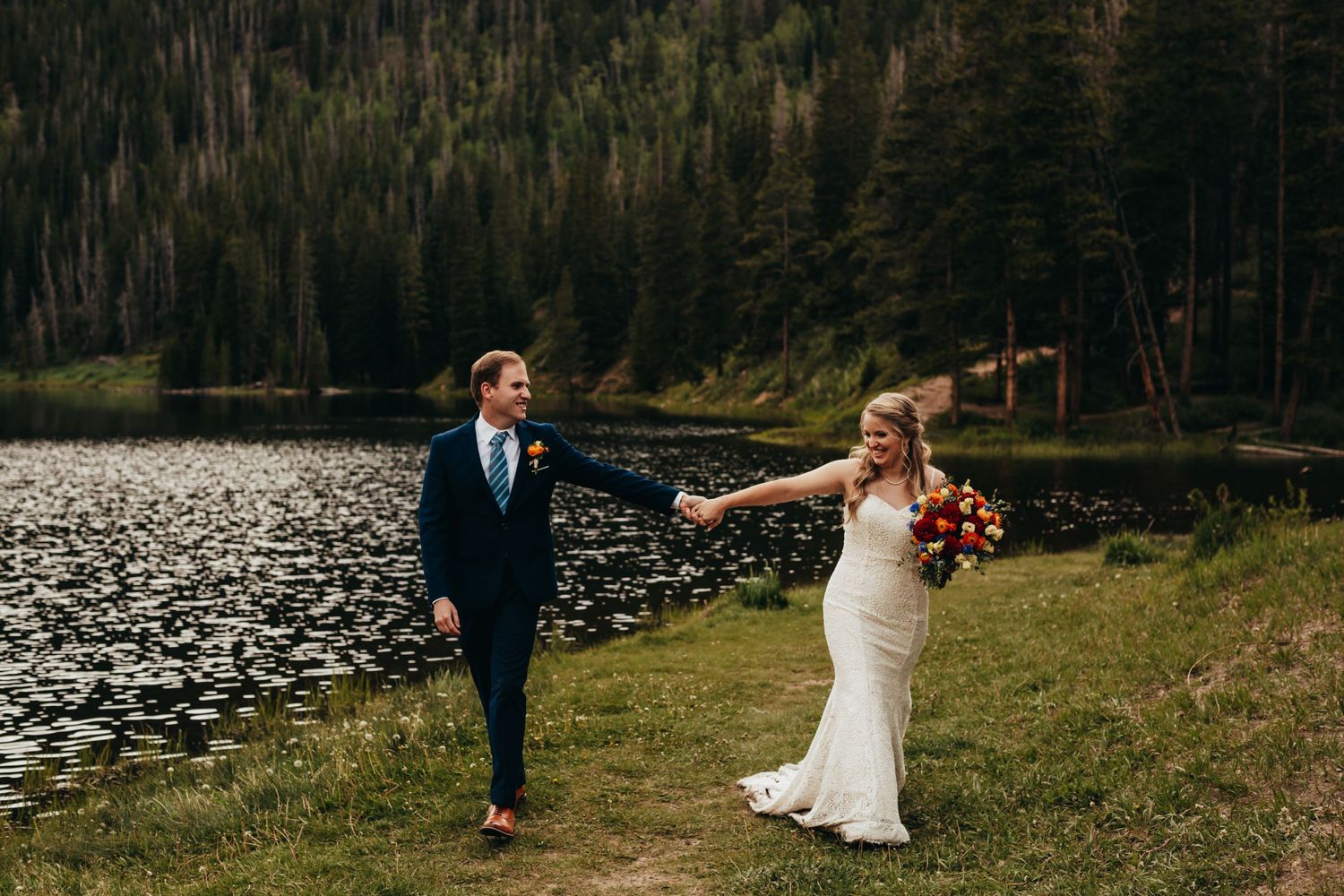 A couple walks hand in hand along a lakeside path in a mountain setting.