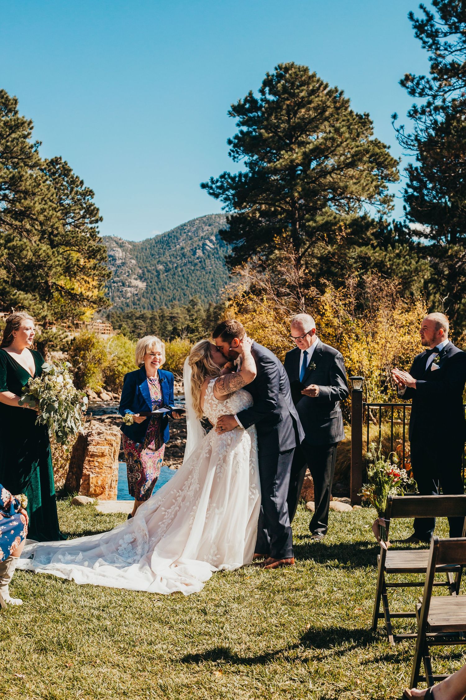 A wedding ceremony takes place outdoors with mountain views and tall pine trees in the background.
