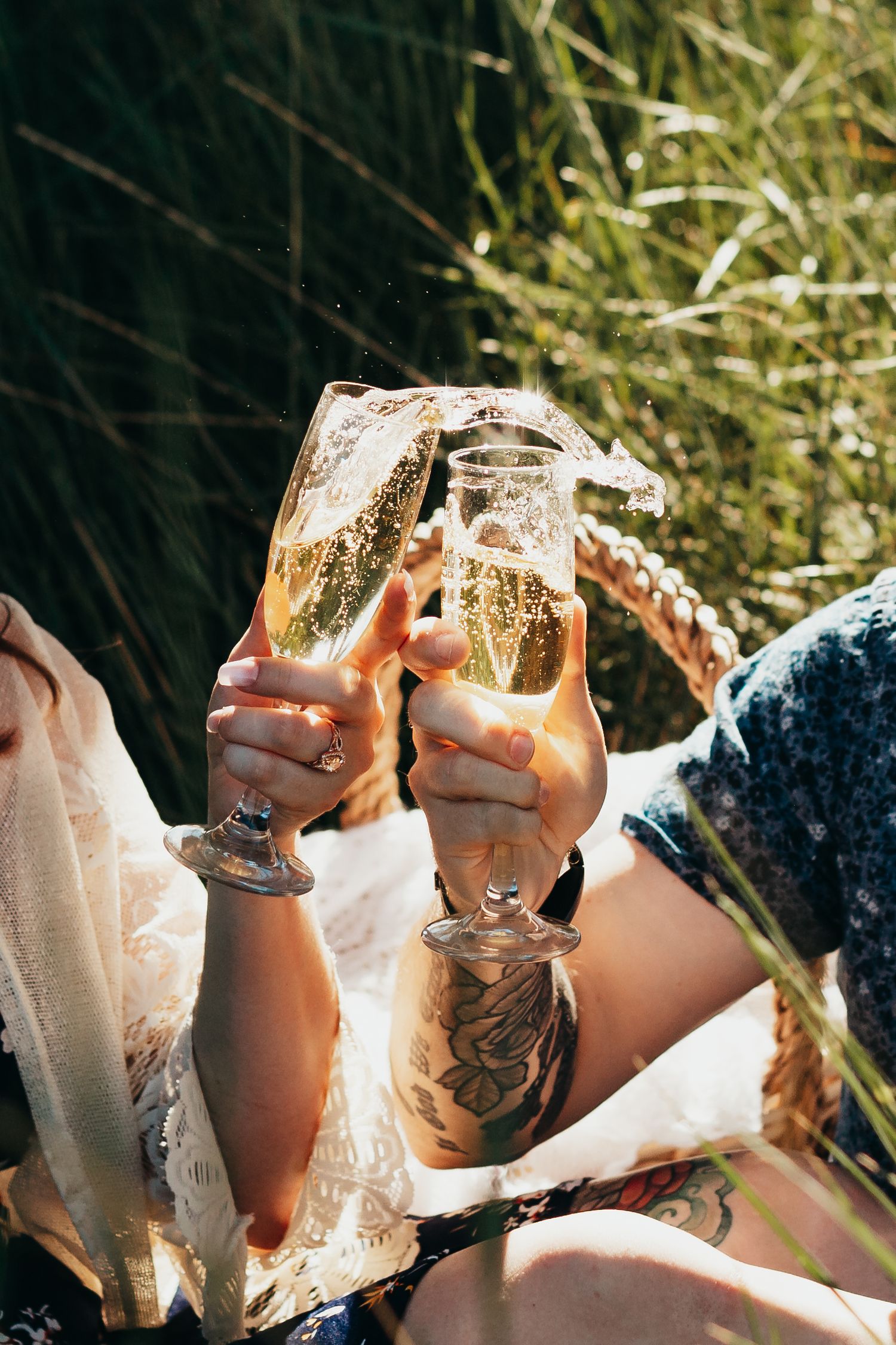 Two hands clinking champagne glasses outdoors with sun flare and grass background.