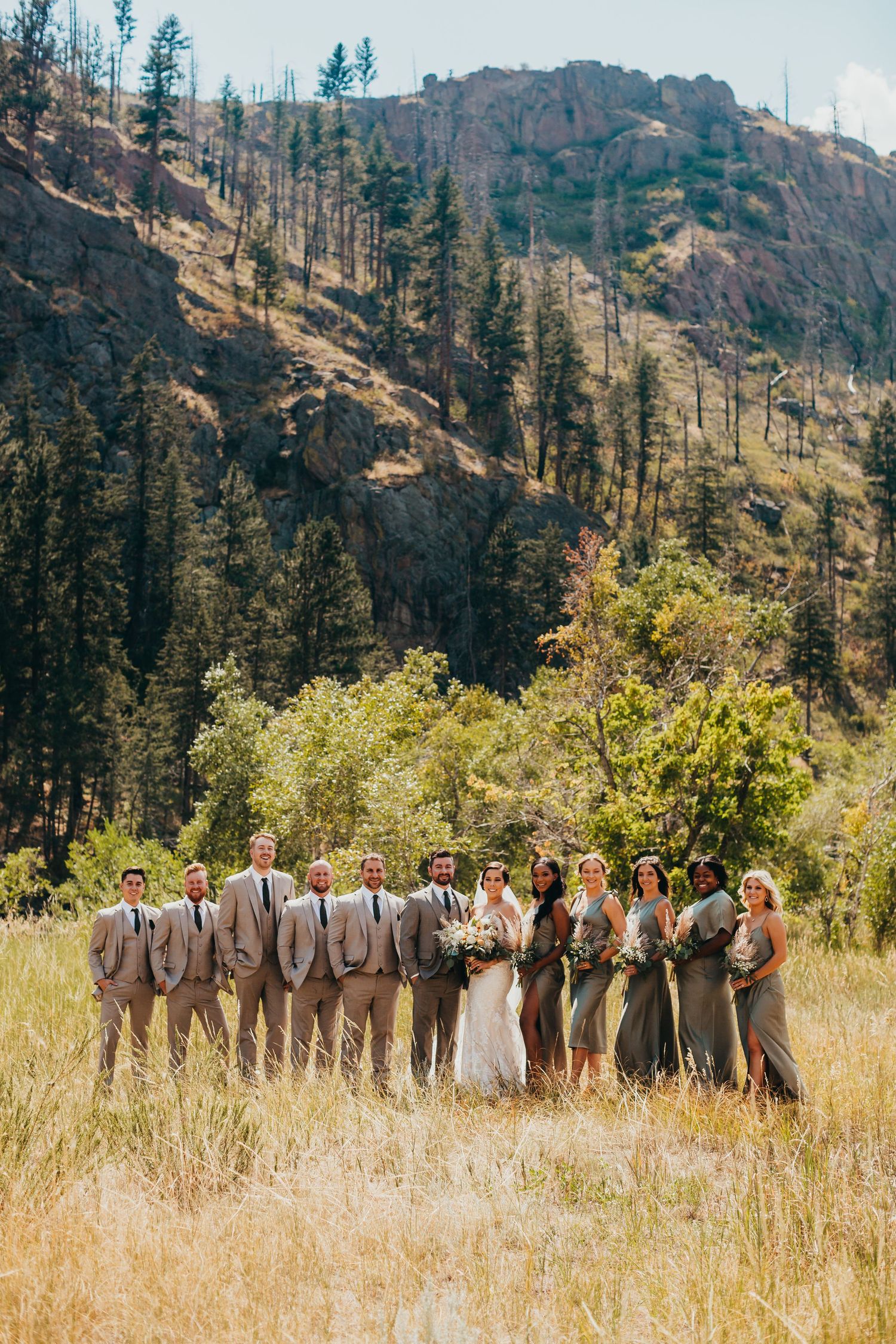 Wedding party in neutral attire poses together in mountain field with dramatic rocky cliffs behind them.