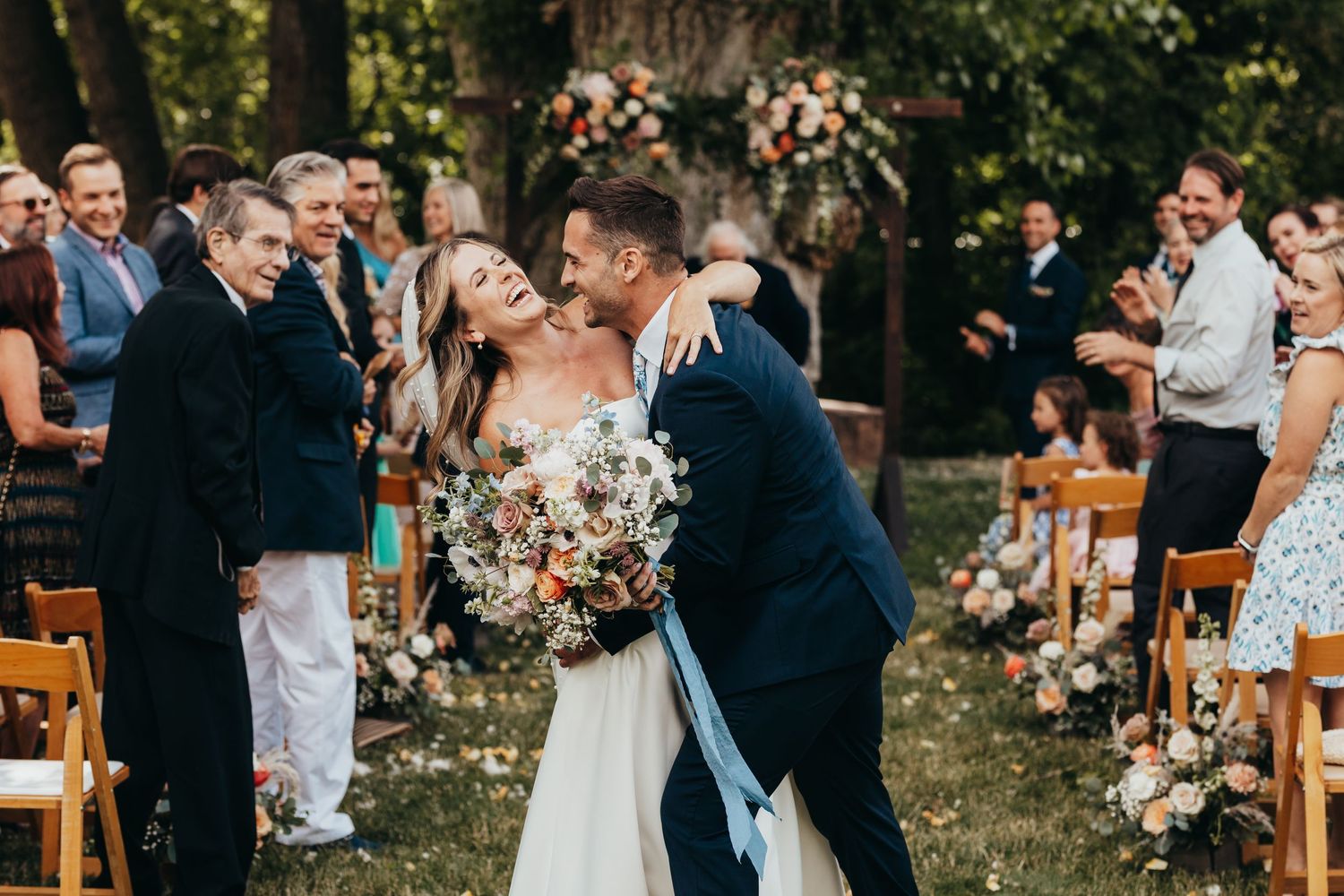 Wedding couple shares intimate moment during outdoor ceremony surrounded by wooden chairs and floral decorations.