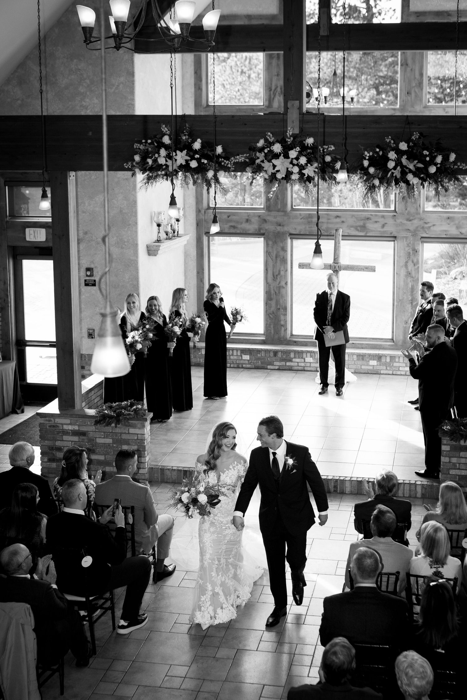Newlyweds walk down aisle in rustic venue with floral decorations overhead.