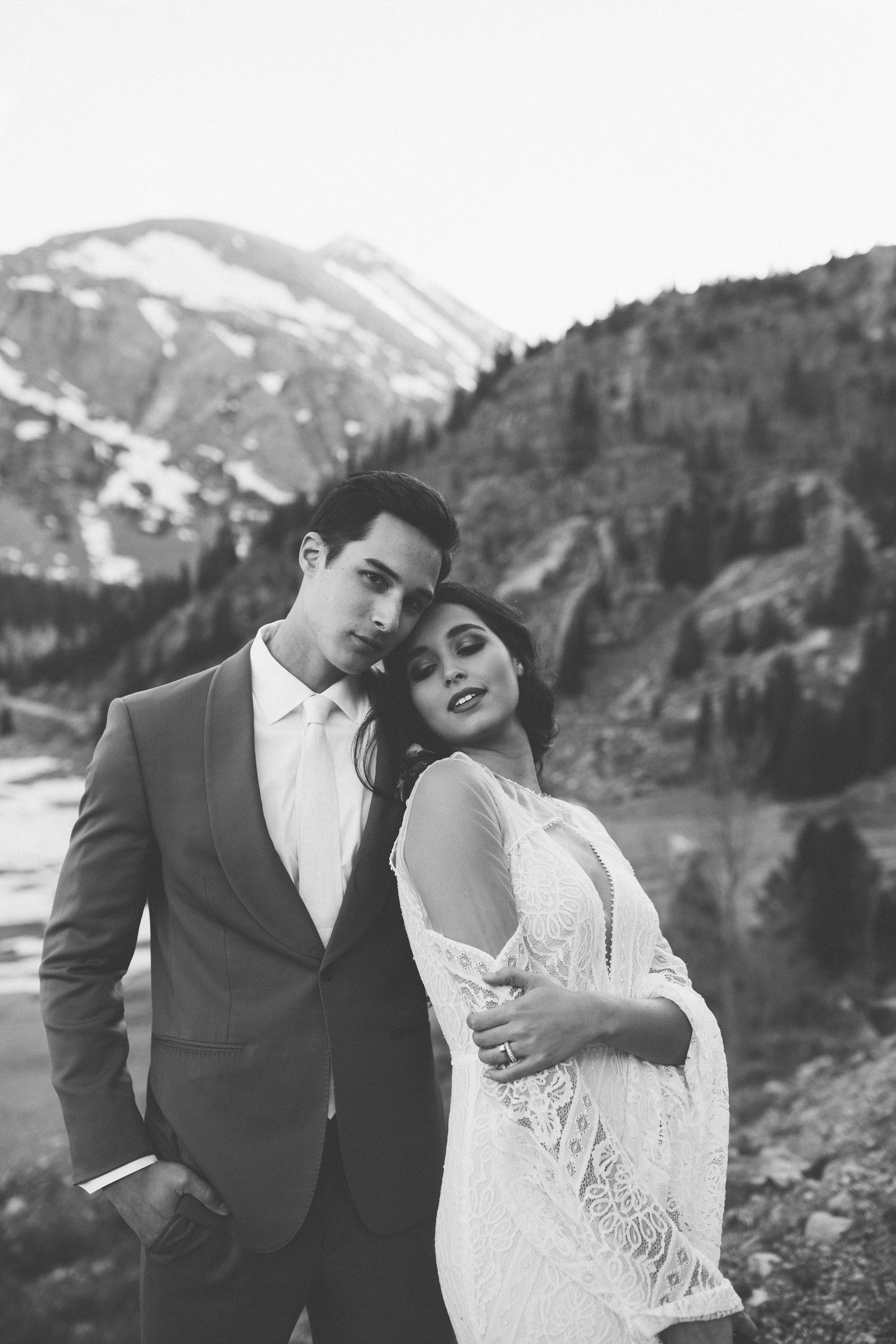 Black and white wedding portrait with mountain peaks and evergreen forest in background.
