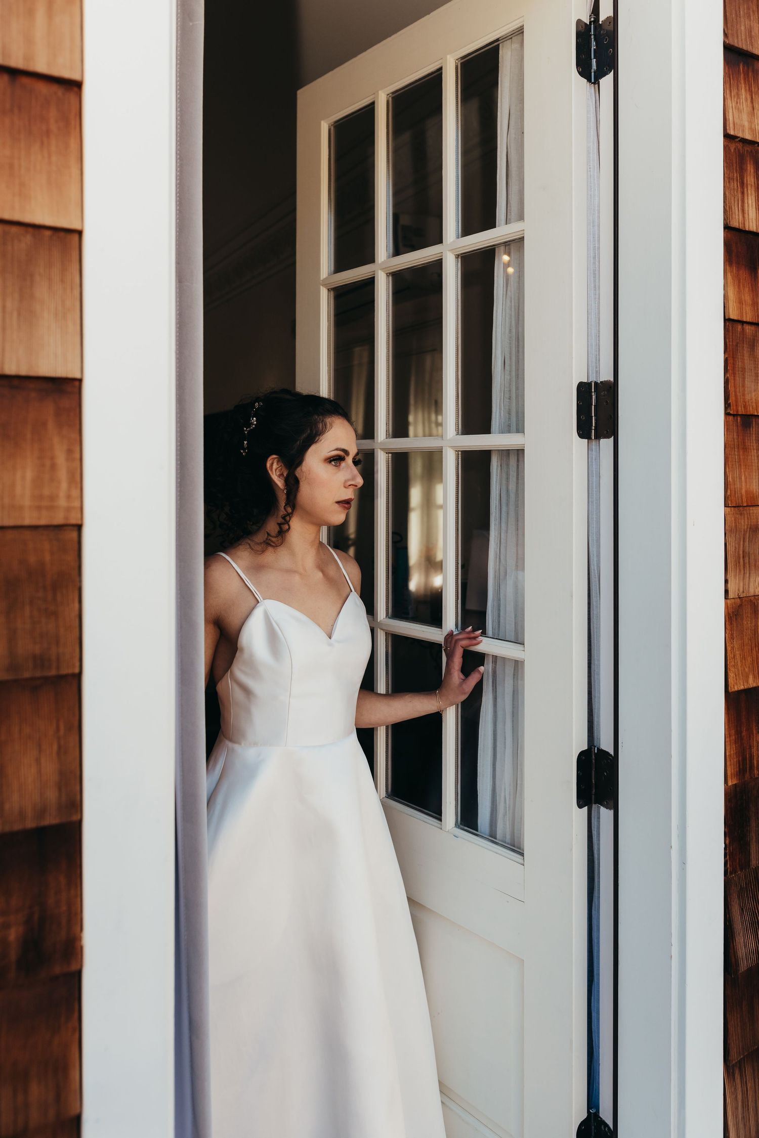 Bride in white dress peers through French door window panes.