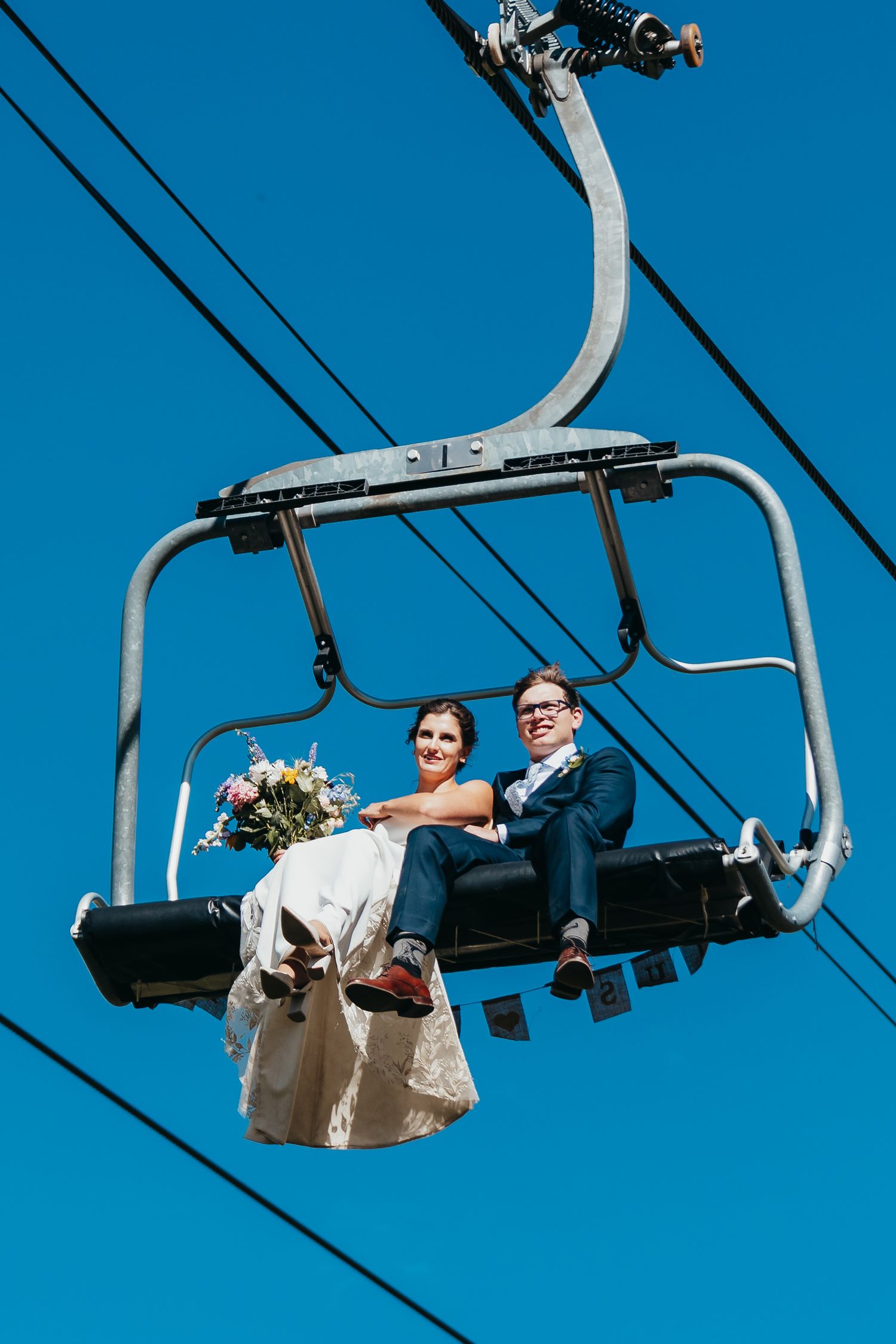 A couple rides a ski lift chair against bright blue sky on their wedding day.