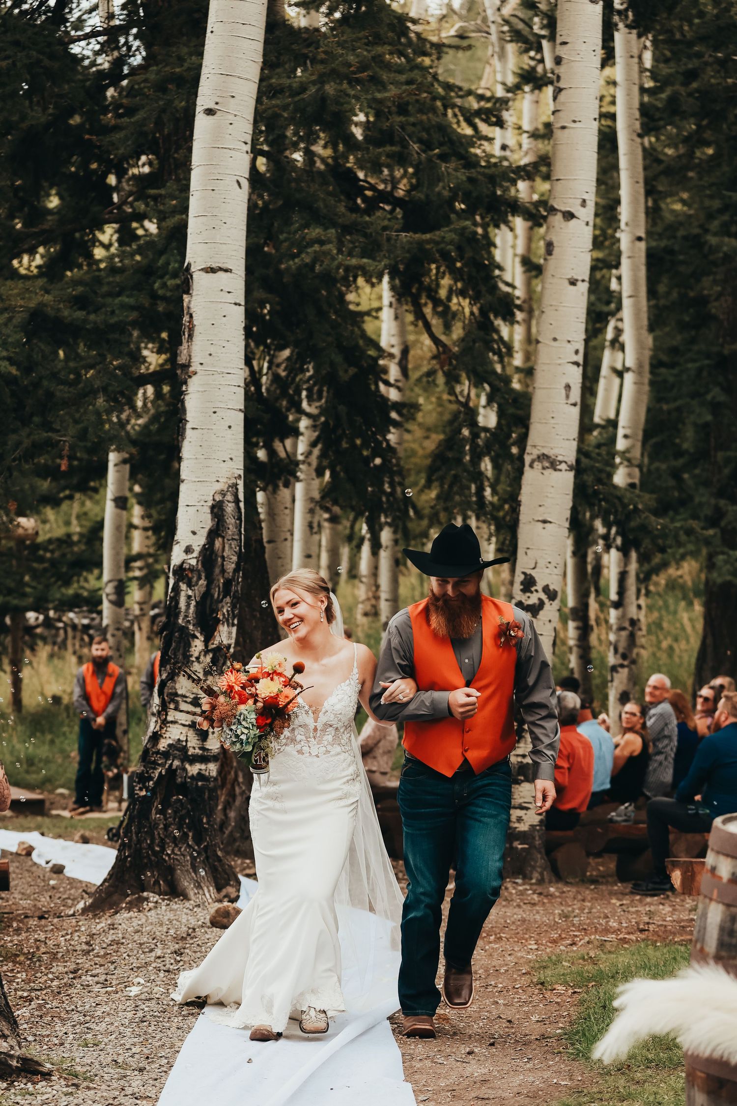 A couple walks down an outdoor aisle lined with aspen trees and wedding guests.
