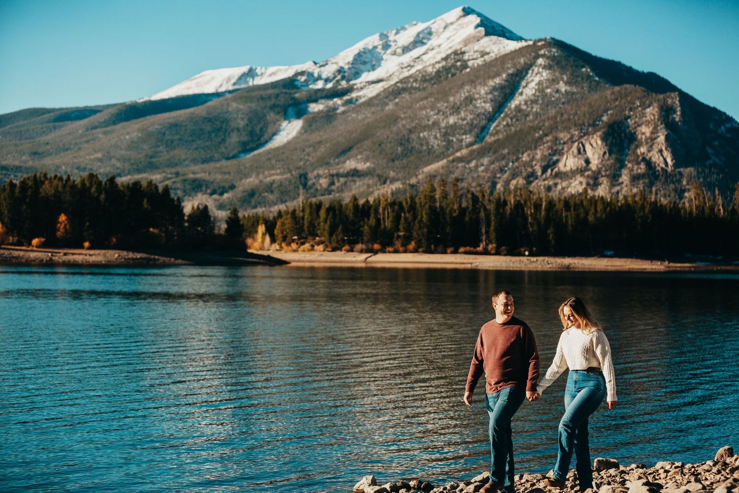 Couple holds hands while walking along mountain lake with snow-capped peaks in background.