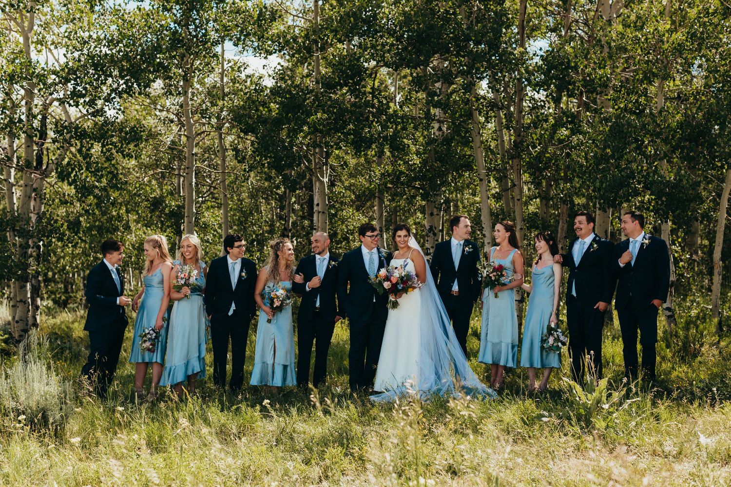 Wedding party in light blue dresses and dark suits poses together against a forest backdrop.