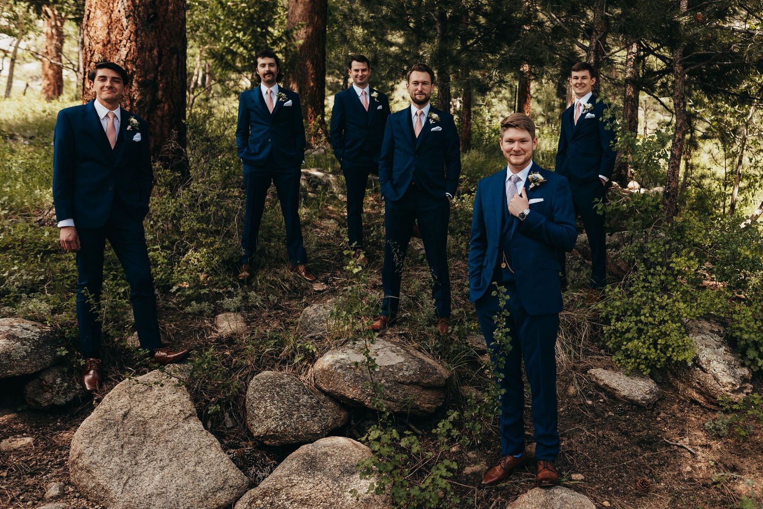 Groomsmen in navy suits pose on rocky terrain among tall pine trees.