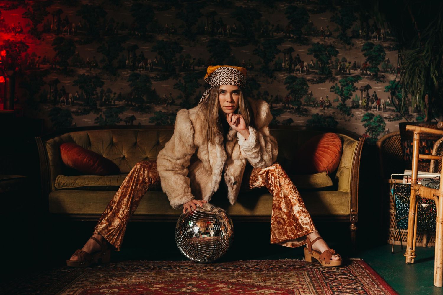 A person in sequined pants poses on a vintage couch with a crystal ball.